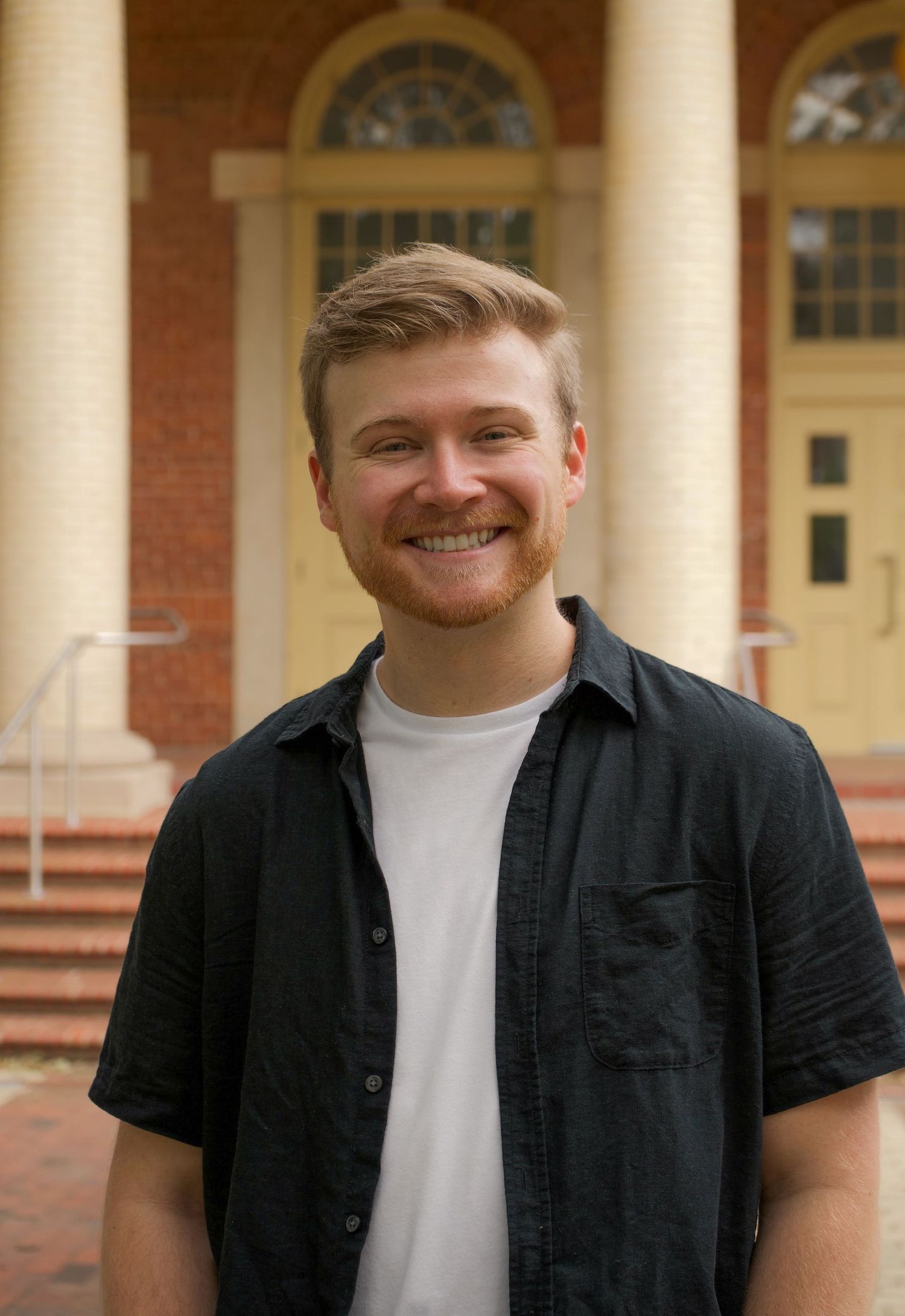 Young man with red beard smiling in front of a brick building with white columns on a NC State campus.