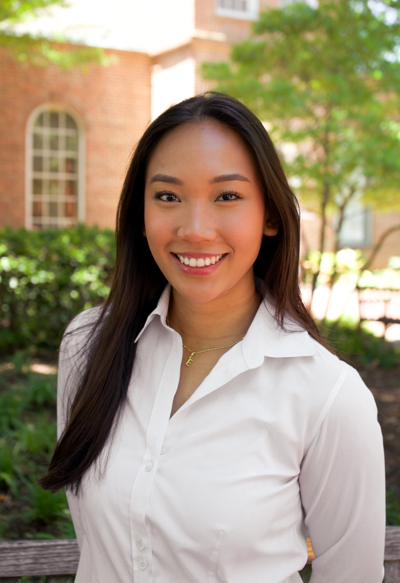 Smiling young woman in white button-up shirt posing outdoors near a brick building with greenery.