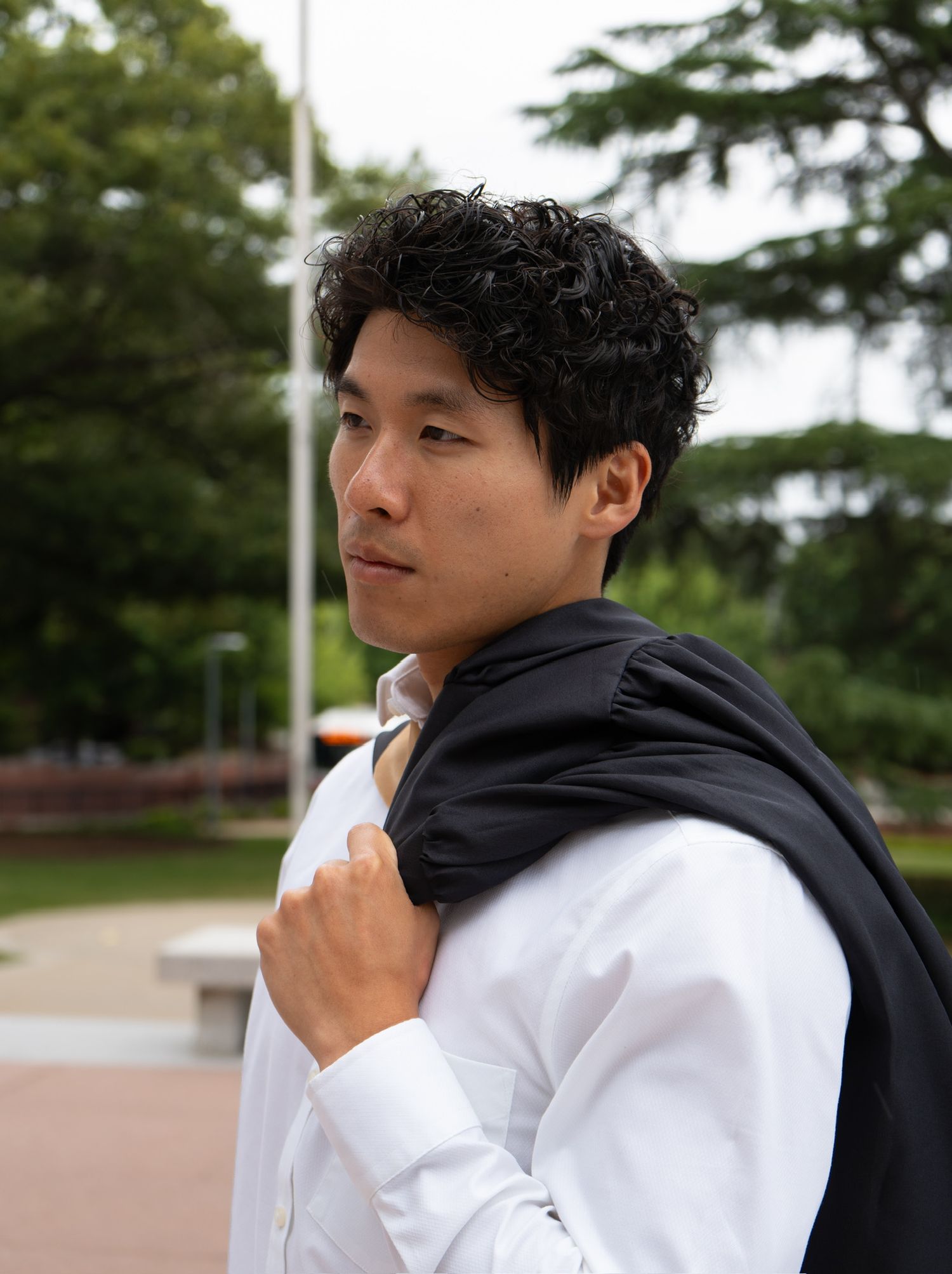 Young man in white shirt carries black graduation gown over shoulder on college campus outdoors.