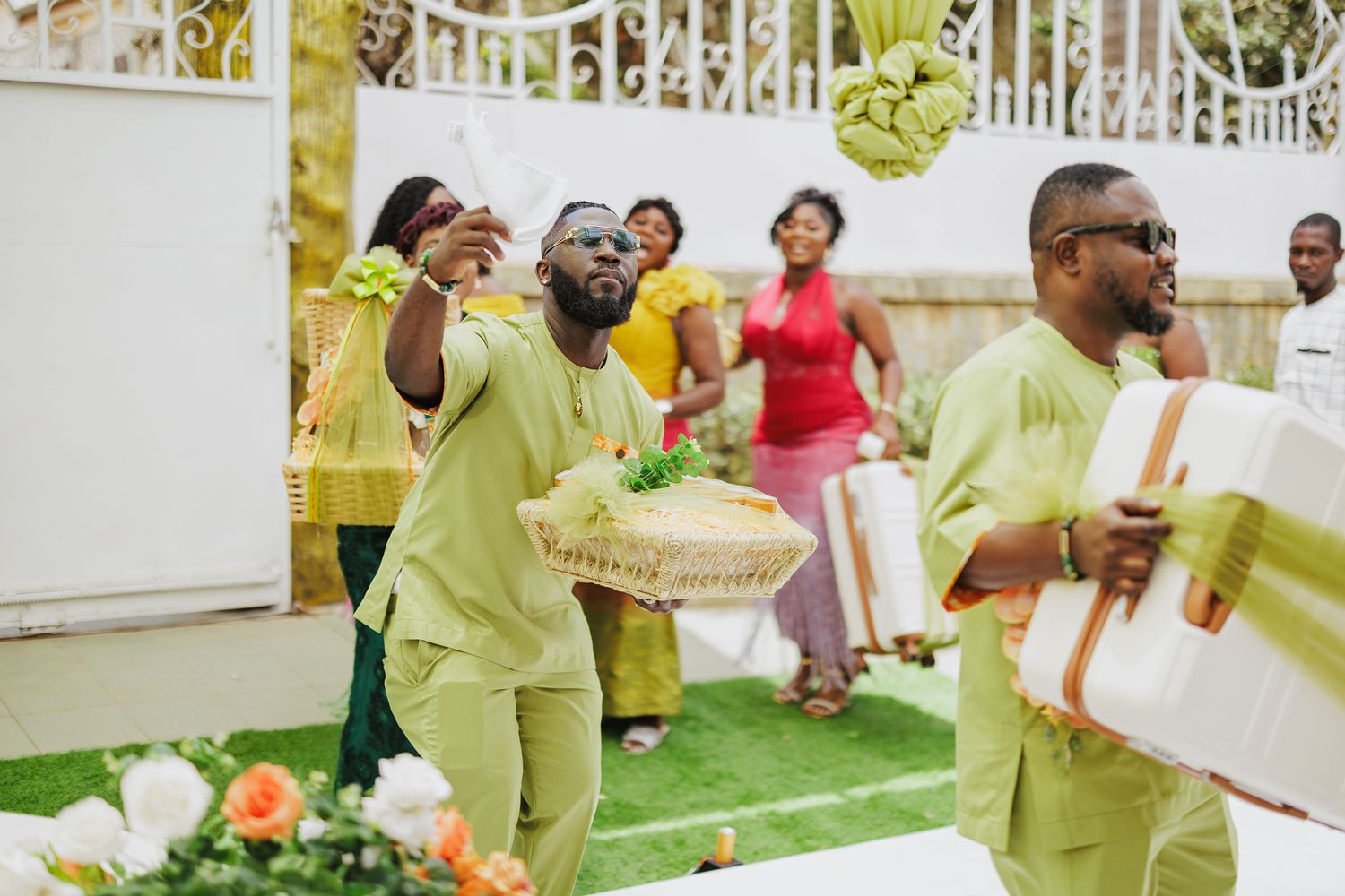 Men in matching green traditional African attire carry gifts and baskets during a vibrant outdoor wedding ceremony.