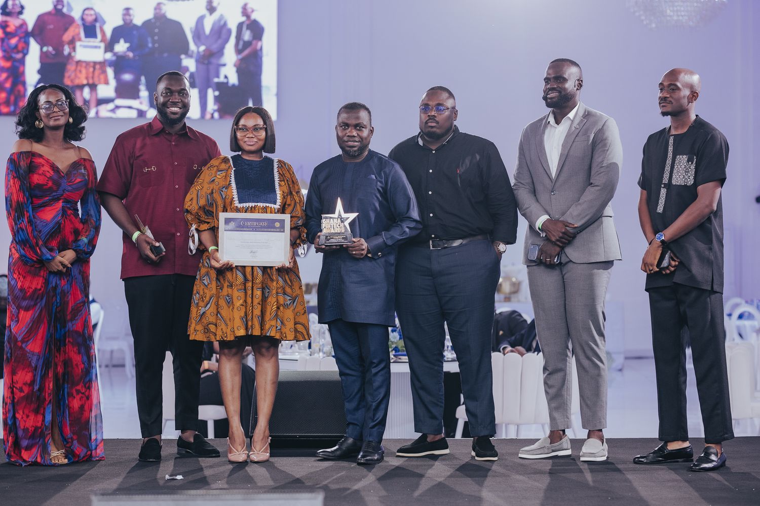 Group of seven people posing together on stage holding award certificate and trophy at gala.