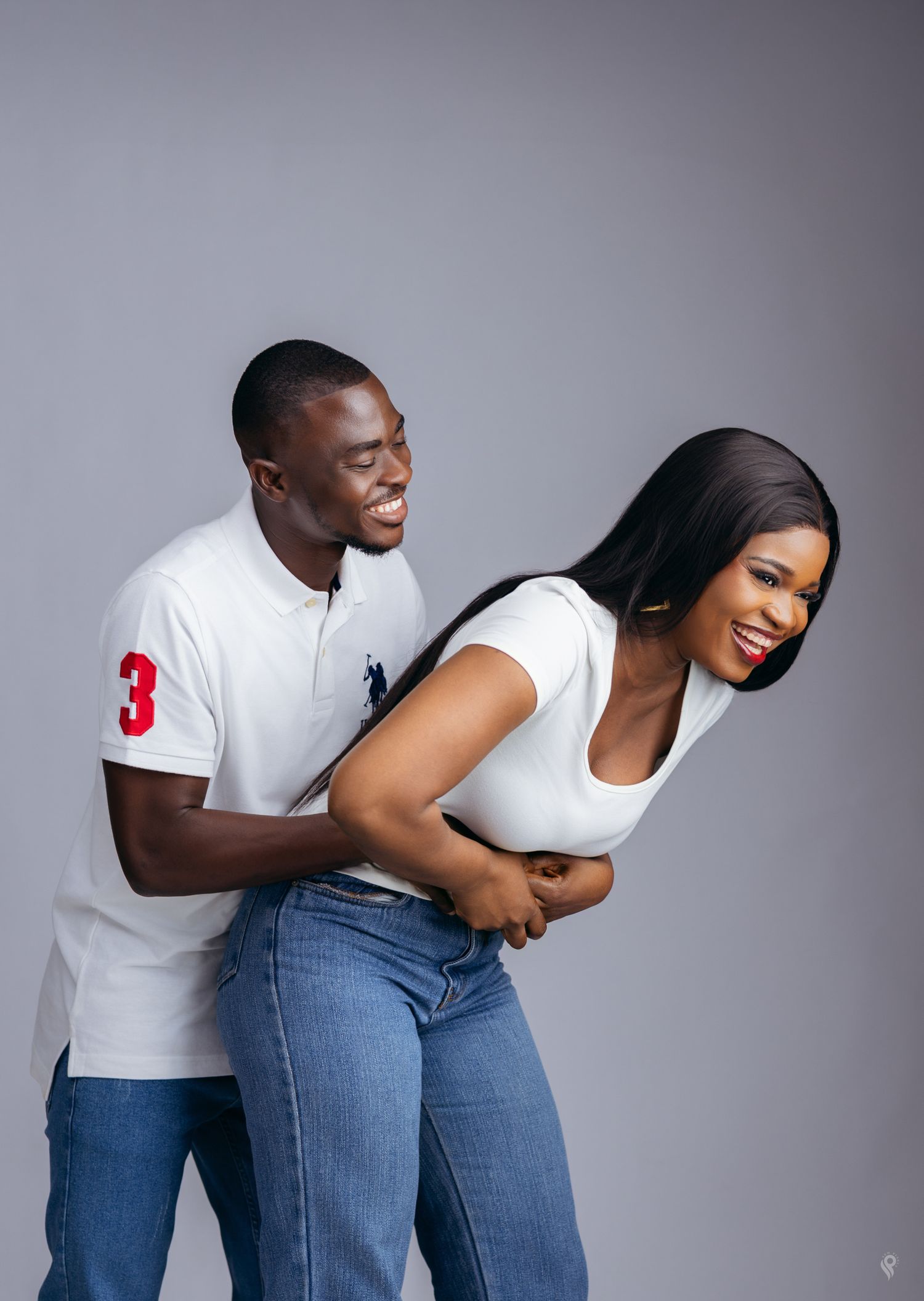 A laughing couple in matching white polo shirts and jeans share a playful moment during a photo shoot.