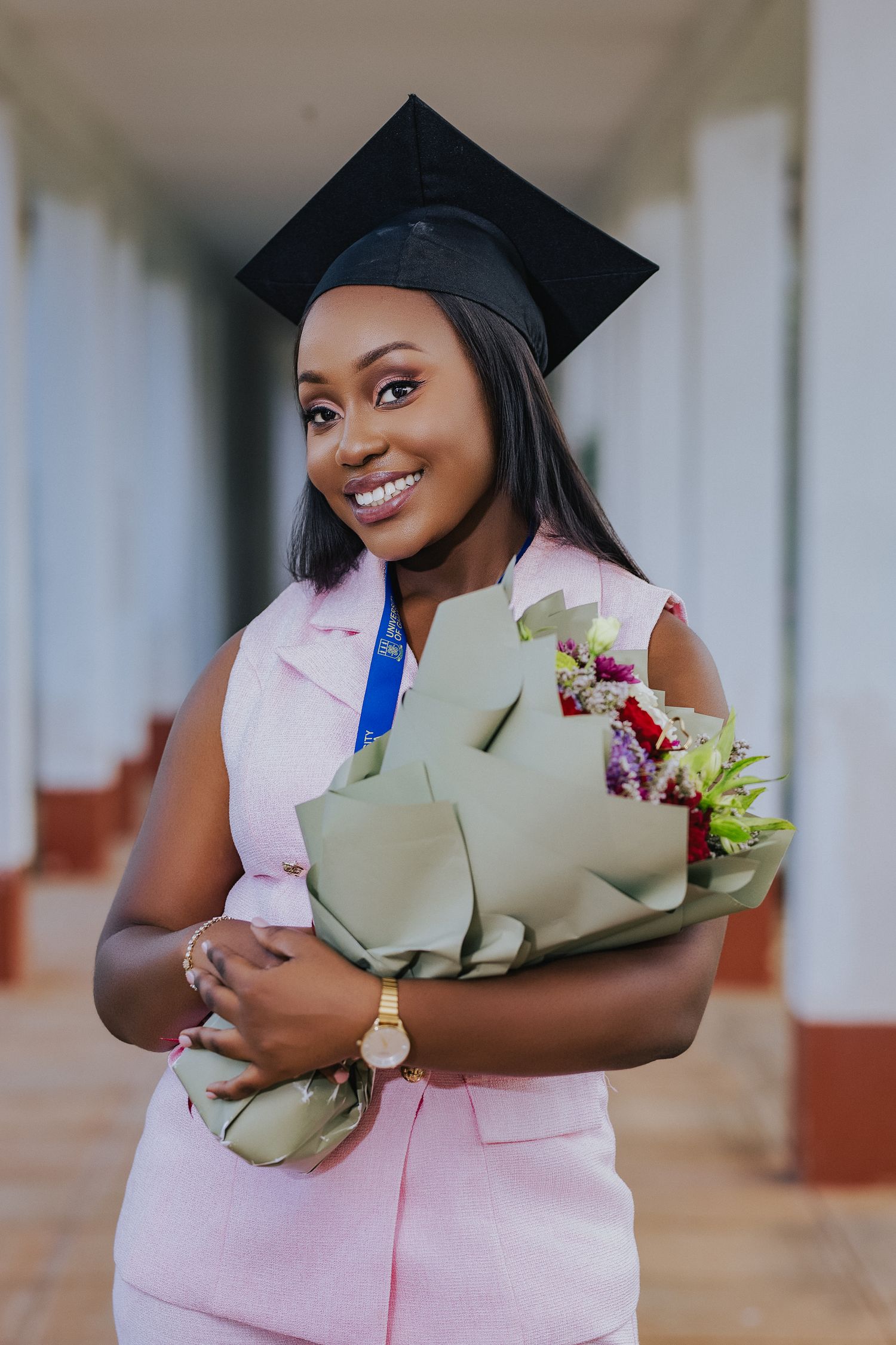 Smiling graduate in pink dress holding flowers with black cap and blue medal.