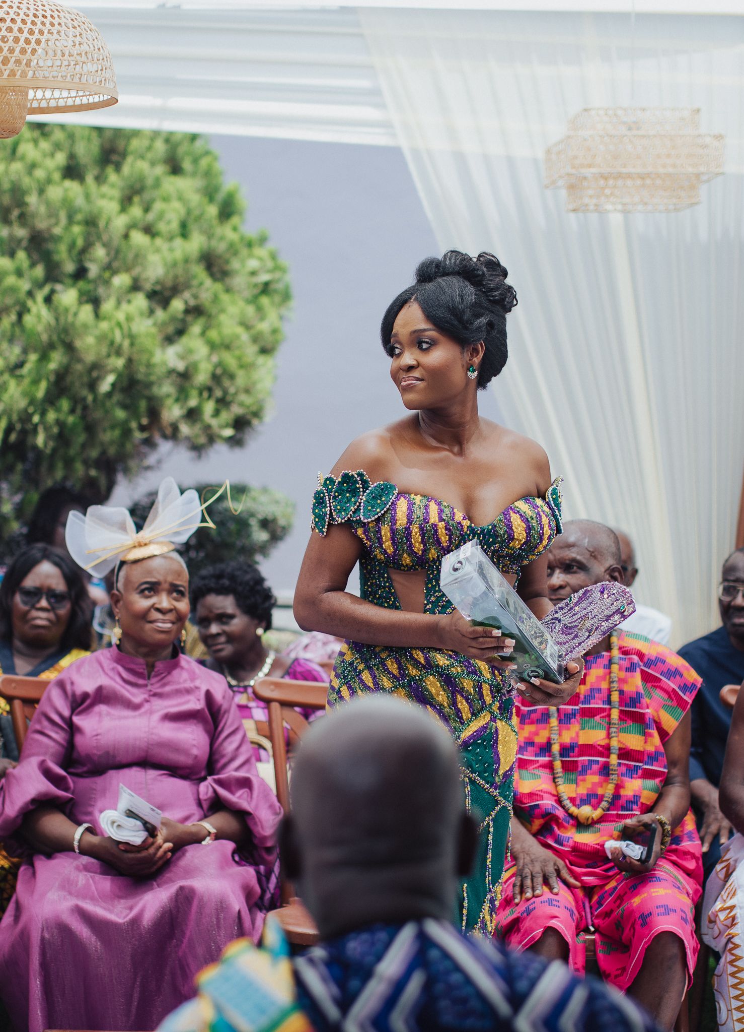 Woman in vibrant Kente off-shoulder dress standing at traditional Ghanaian outdoor wedding ceremony.