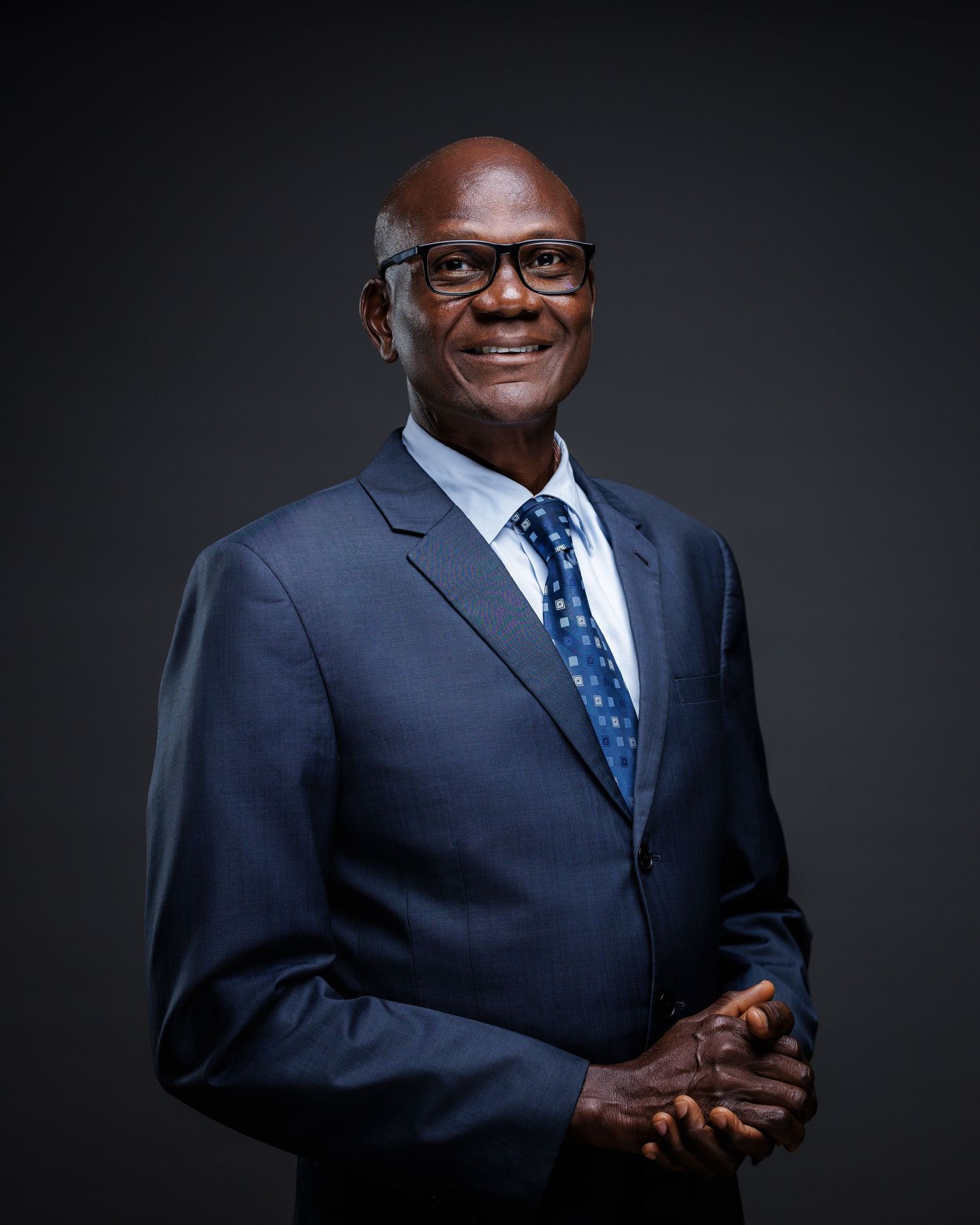 Elderly man in blue suit and glasses smiling warmly in professional corporate portrait.