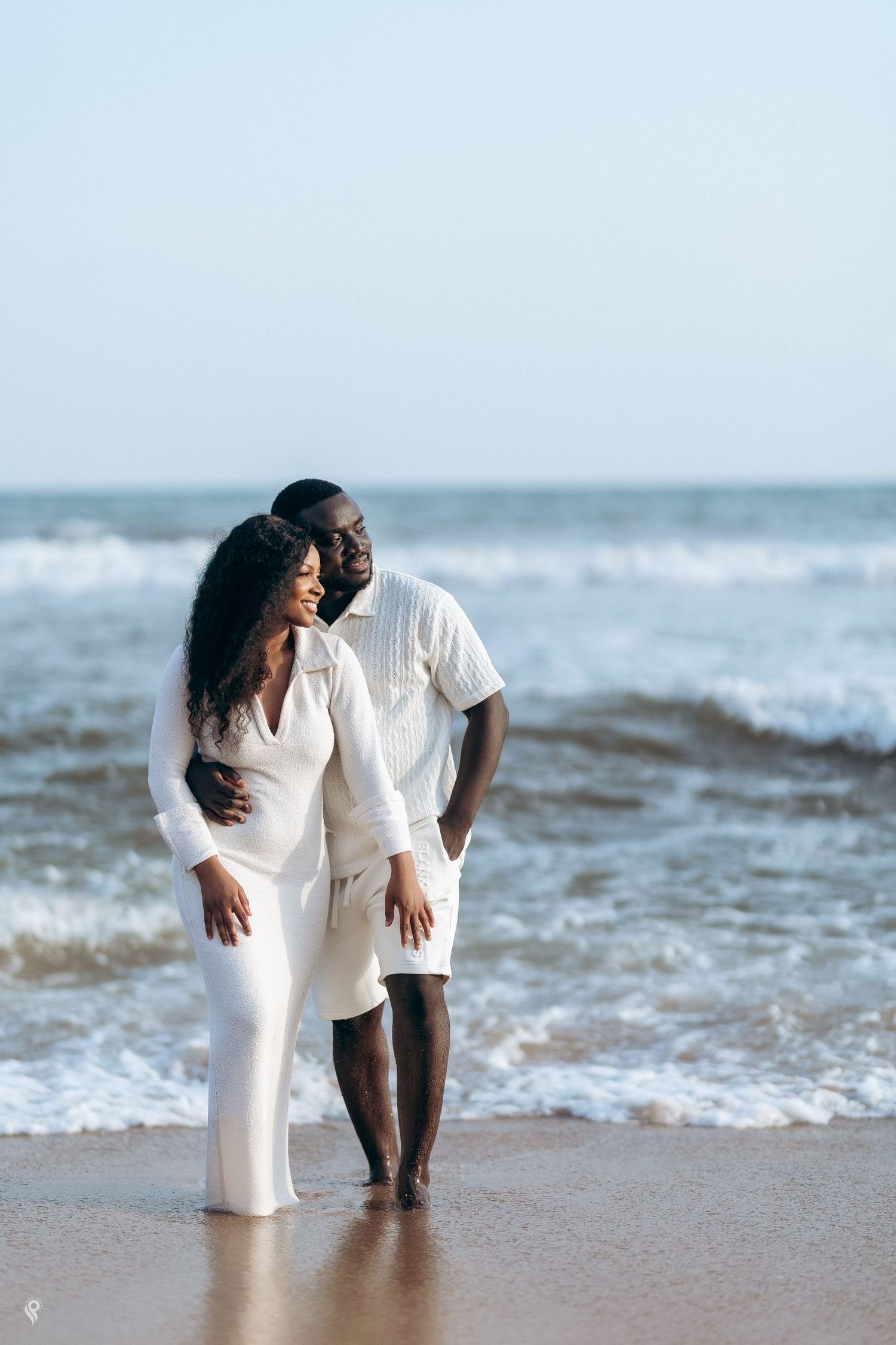 Romantic Black couple in white outfits embracing on a sandy beach with ocean waves behind them.