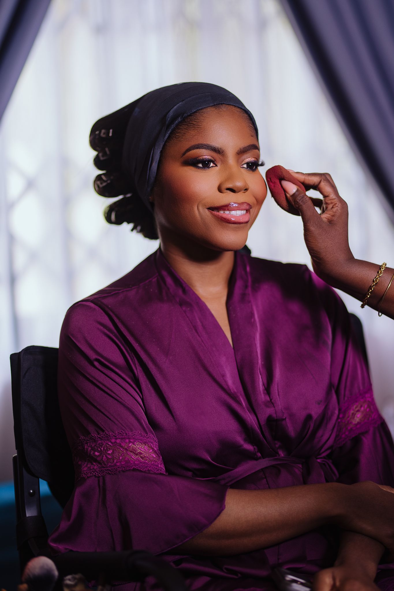 A bride in a purple satin robe gets makeup applied while wearing a black head wrap.