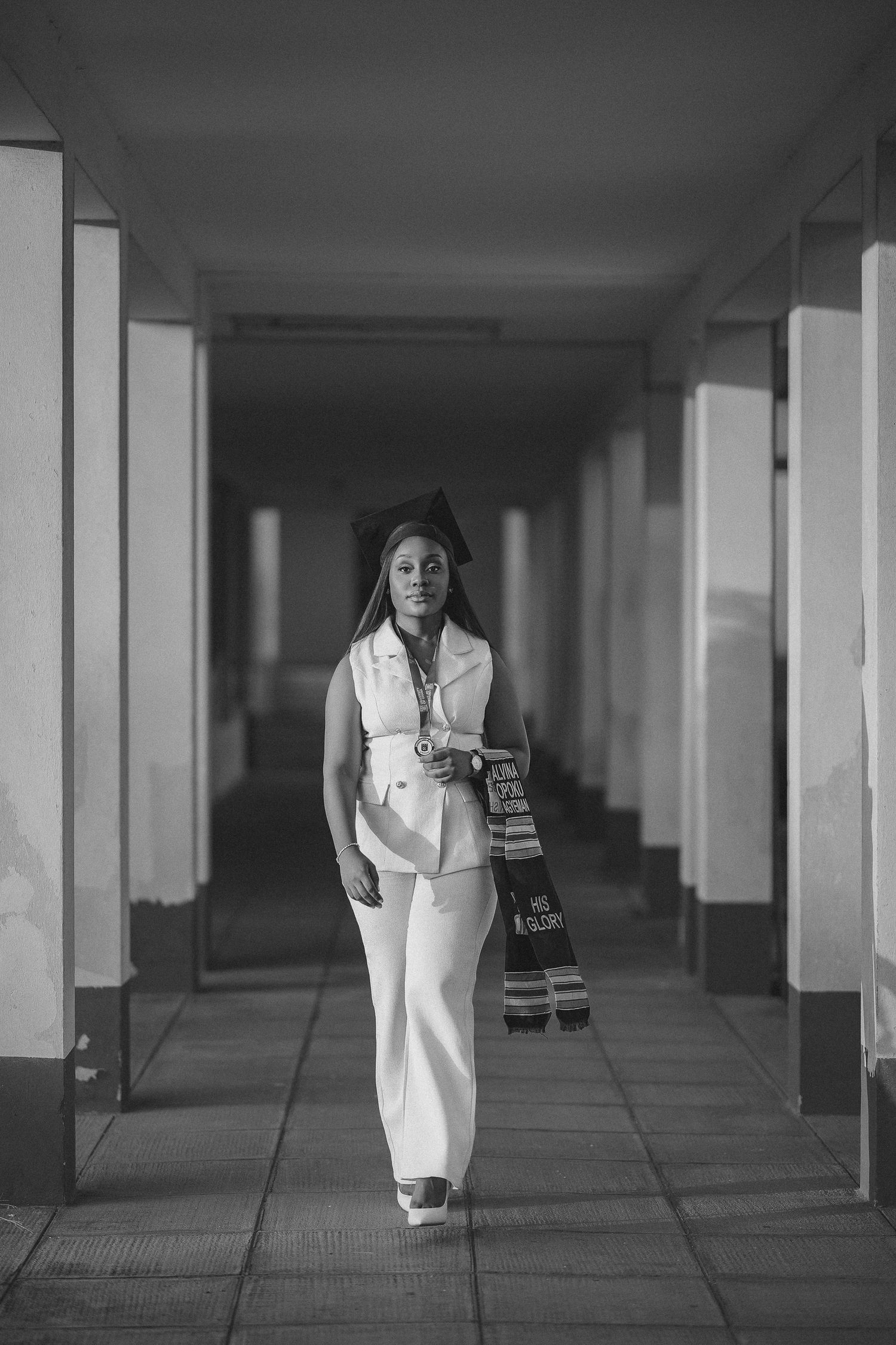 Black and white photo of a graduate in white suit and cap walking through a long corridor.