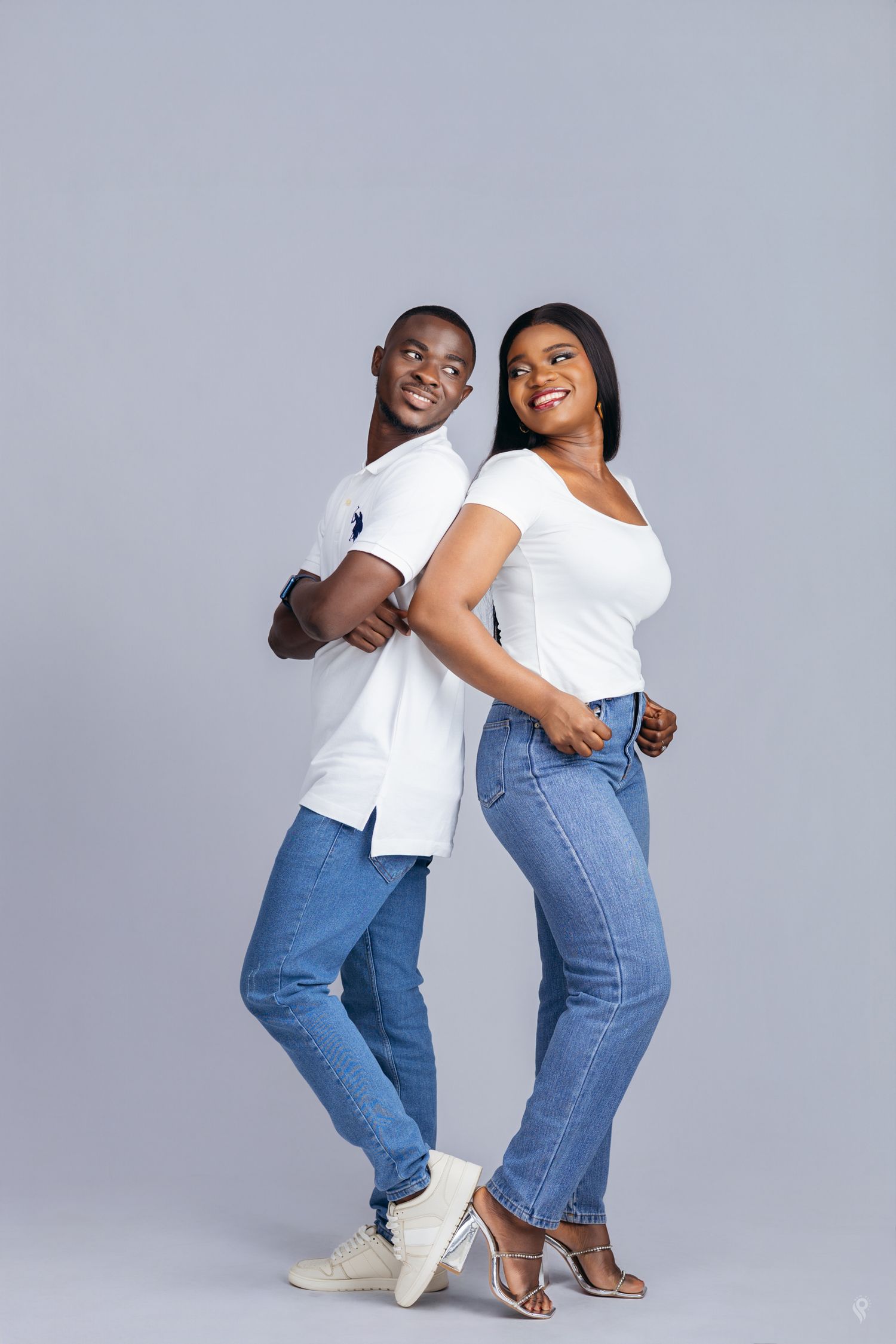 Stylish Black couple in matching white tops and blue jeans posing back-to-back on gray background.
