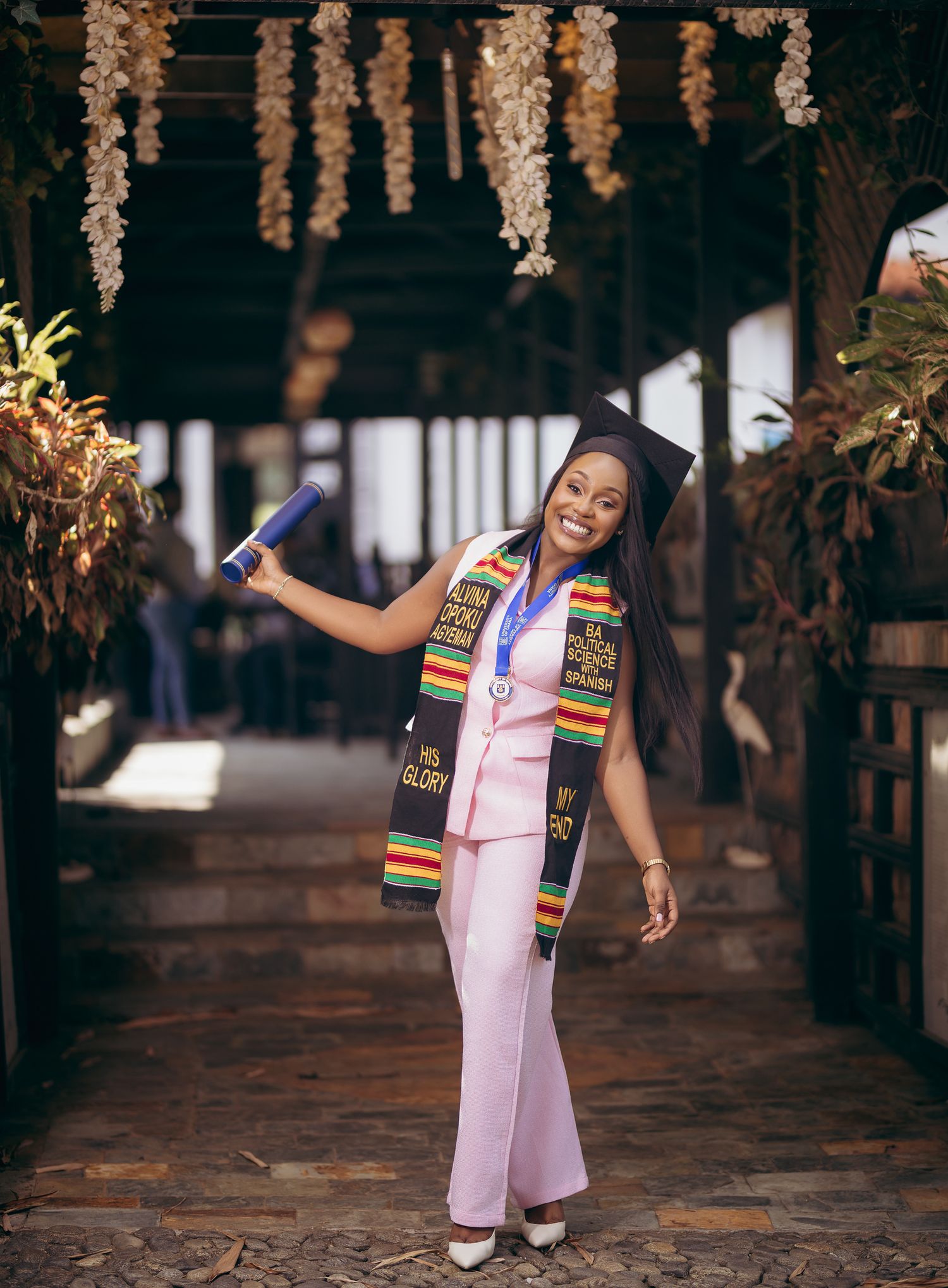 Graduate in pink pantsuit and cap walking confidently under floral archway holding diploma.