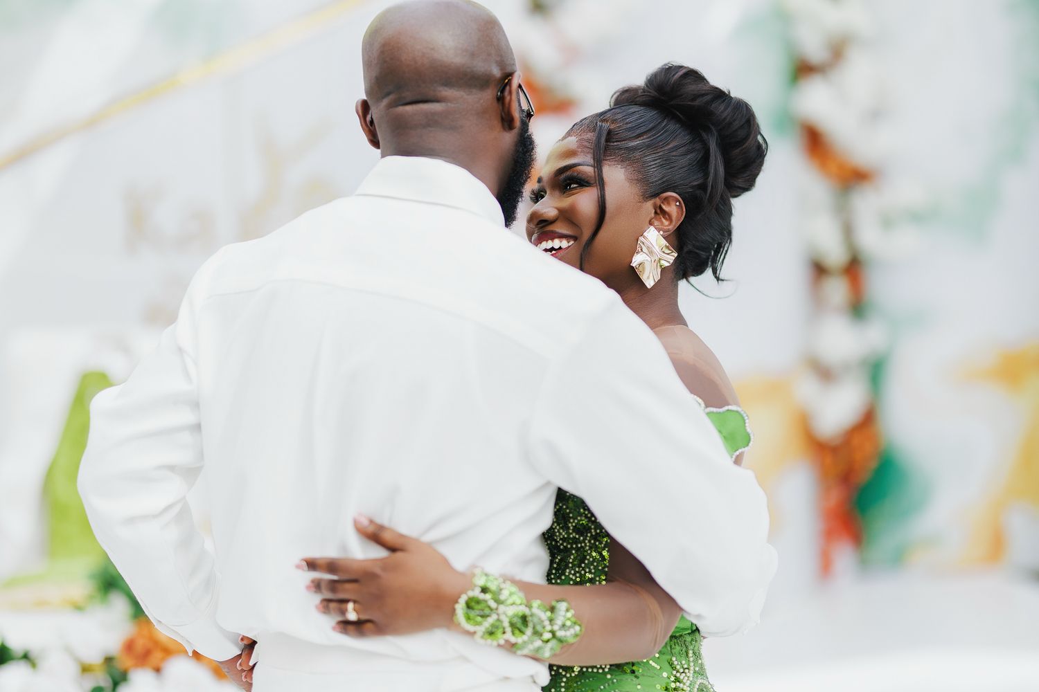 Smiling bride in green dress embraces groom in white suit at outdoor wedding venue.