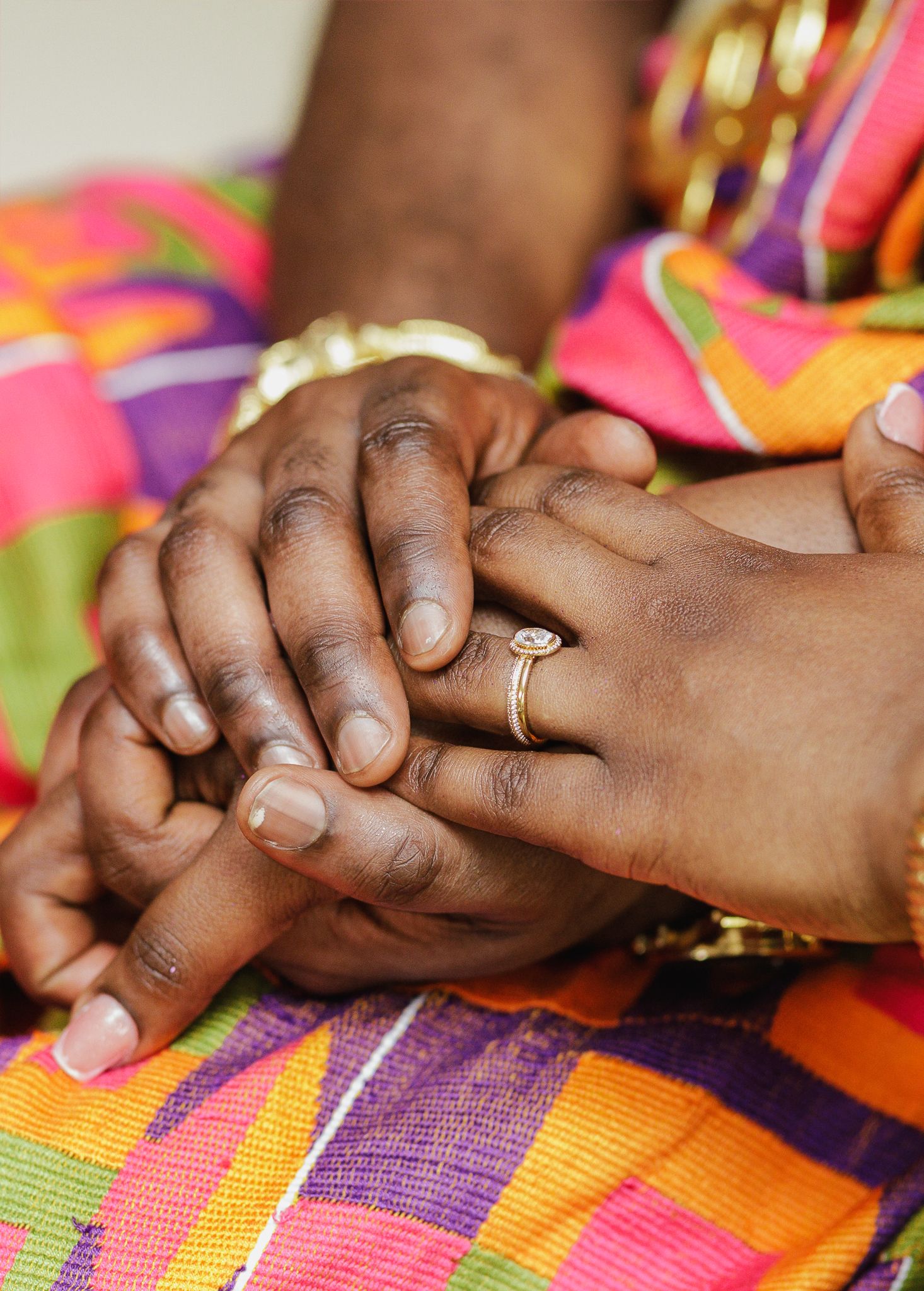 Couple holding hands with engagement ring over colorful Kente fabric.