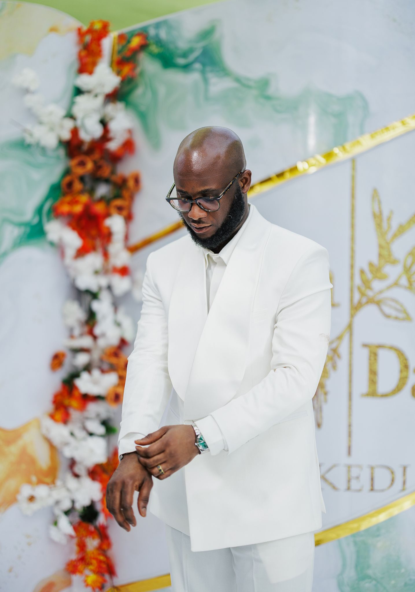 Groom in white suit adjusting cufflinks at a floral wedding backdrop.