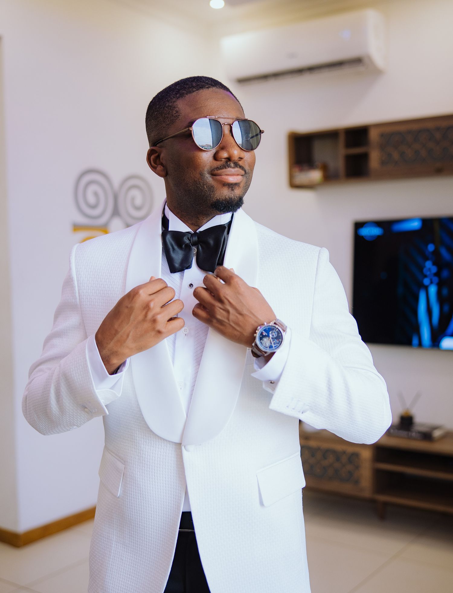 Stylish groom in white tuxedo adjusting bow tie wearing sunglasses and luxury watch indoors.