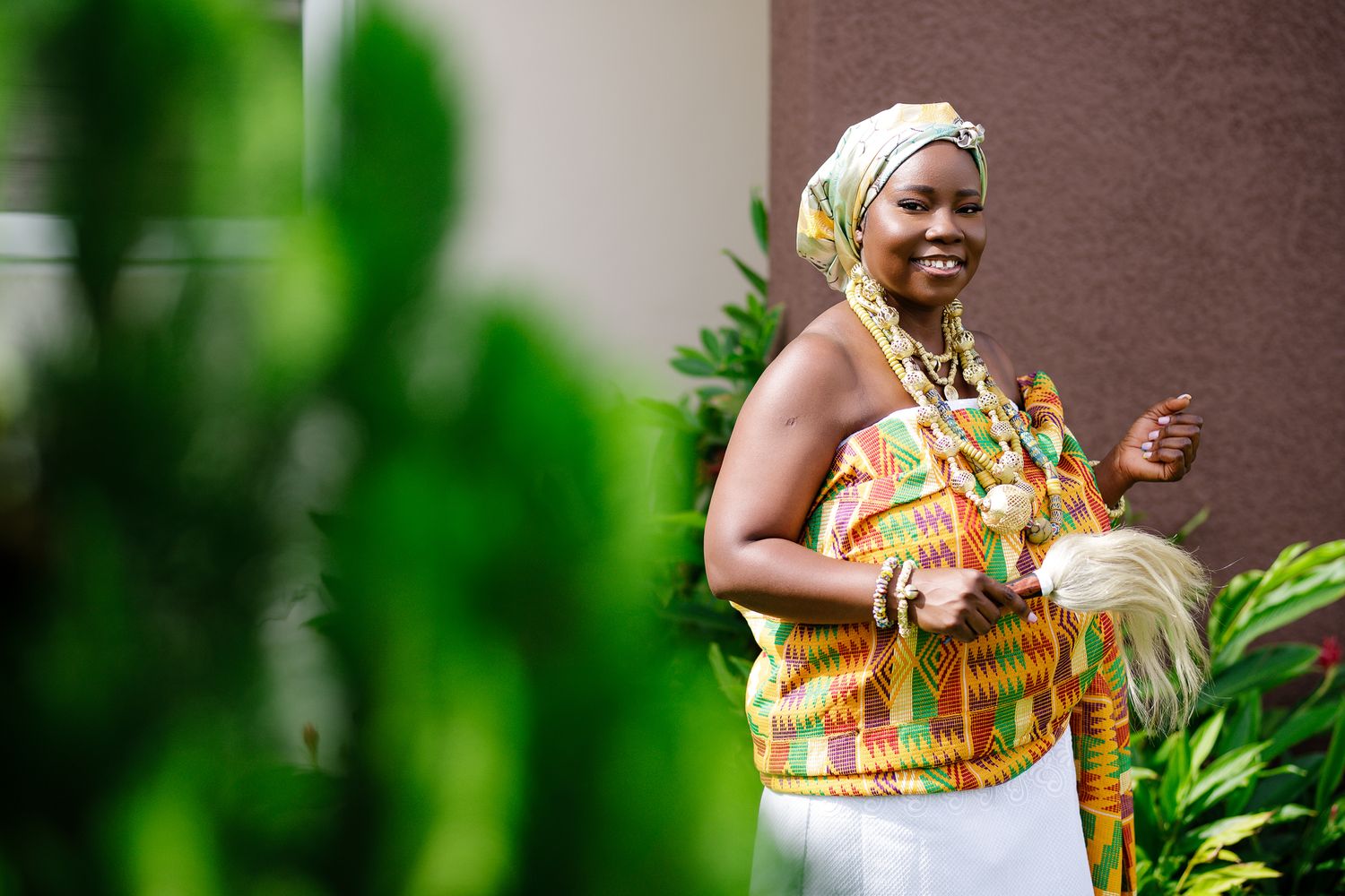 Joyful woman in kente print attire and gold jewelry smiles outdoors during a traditional African celebration.