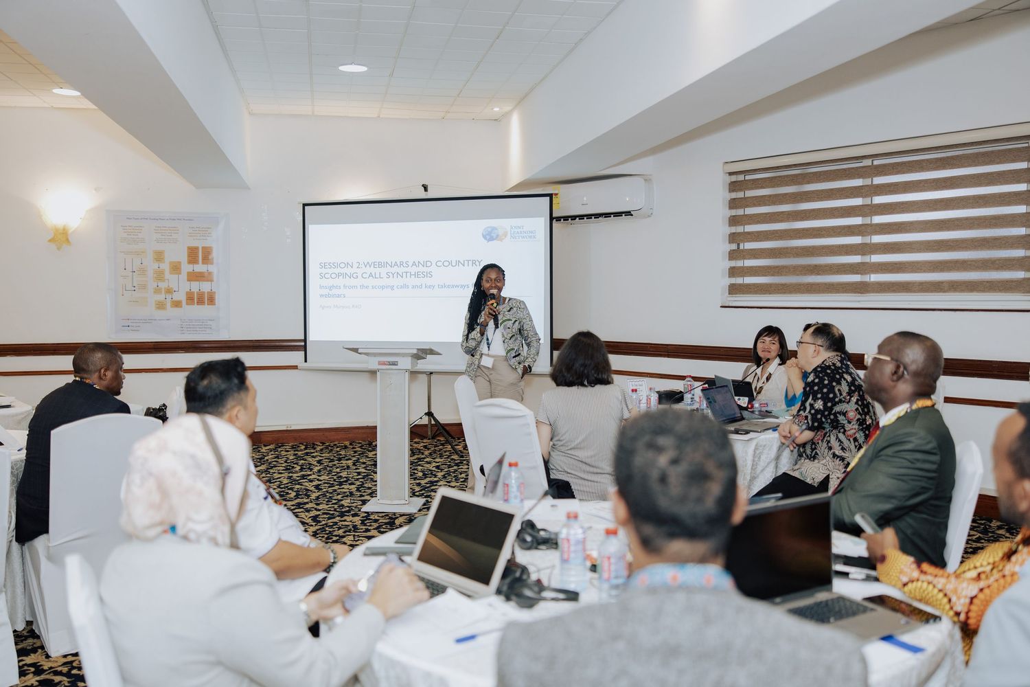 A presenter addresses attendees at a professional workshop in a conference room with a projector screen and laptops.