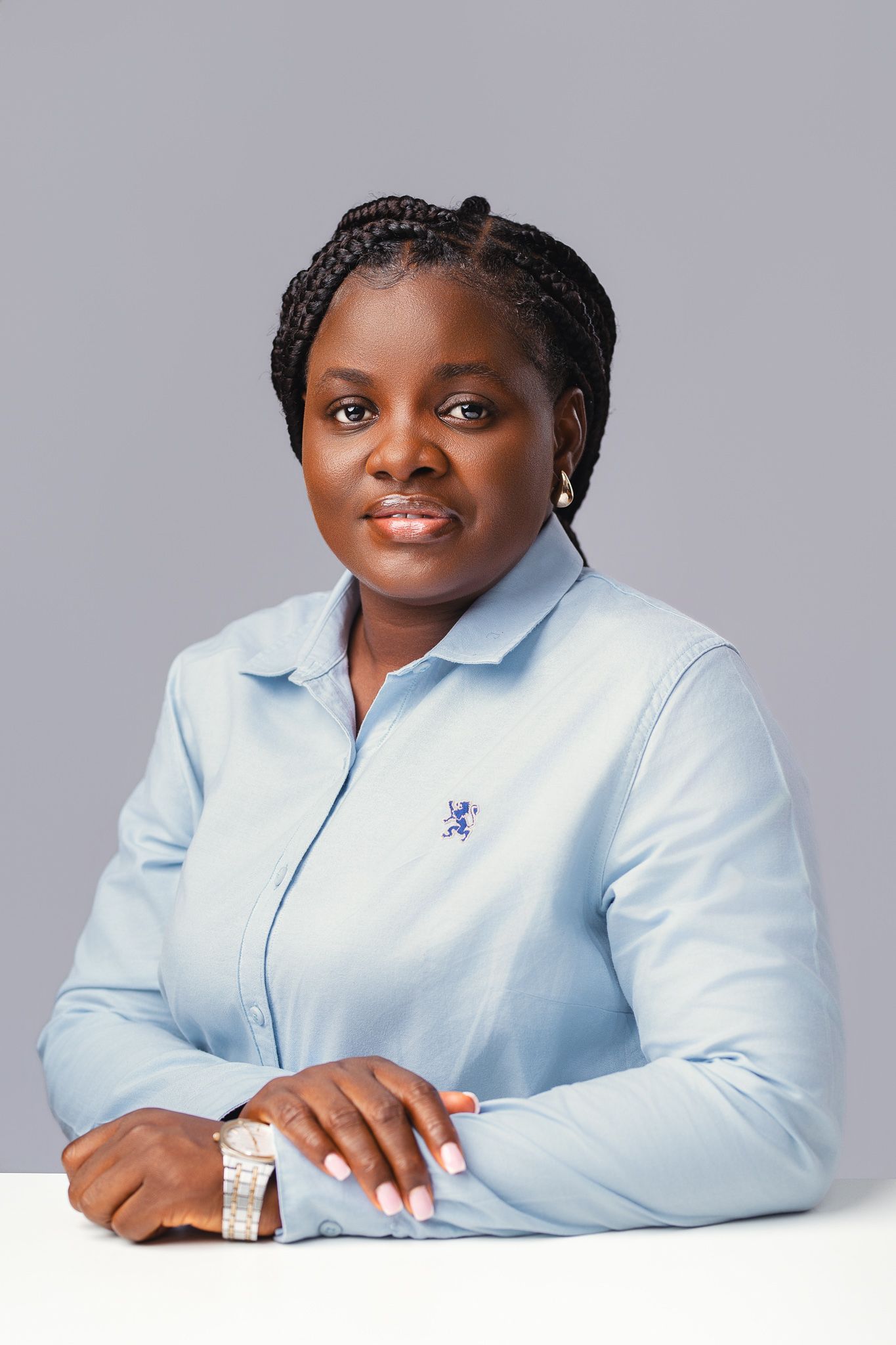 Professional Black woman in blue button-up shirt with braids, seated at white table on gray background.
