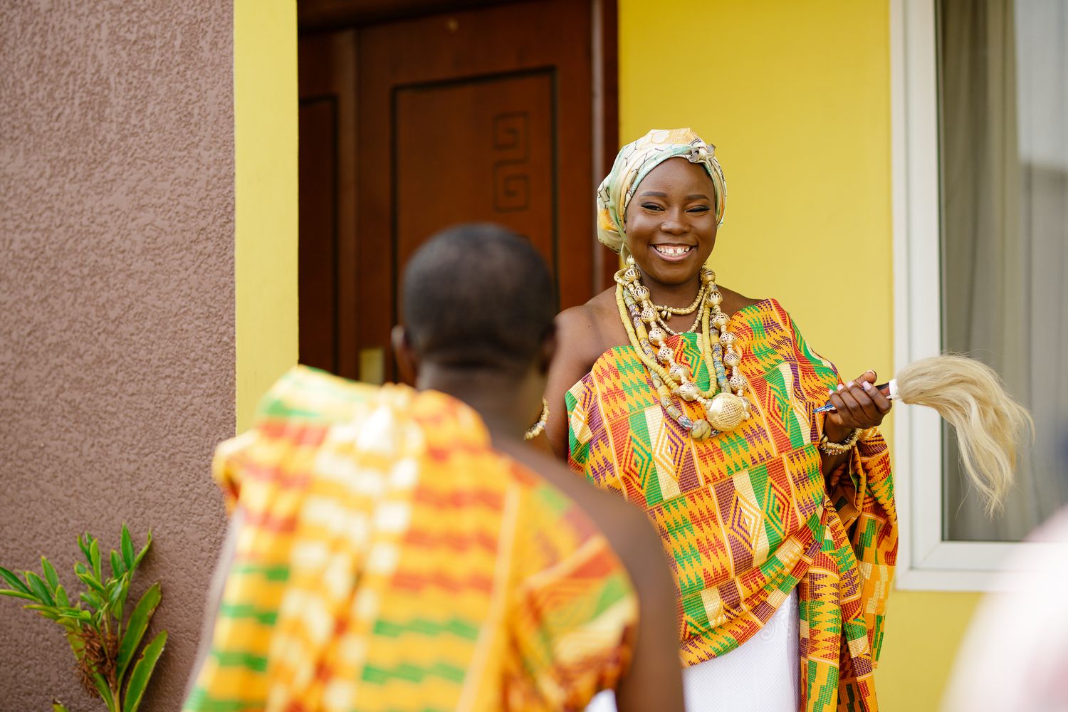 Smiling woman in colorful kente cloth and headwrap holds a fan during a traditional Ghanaian wedding.