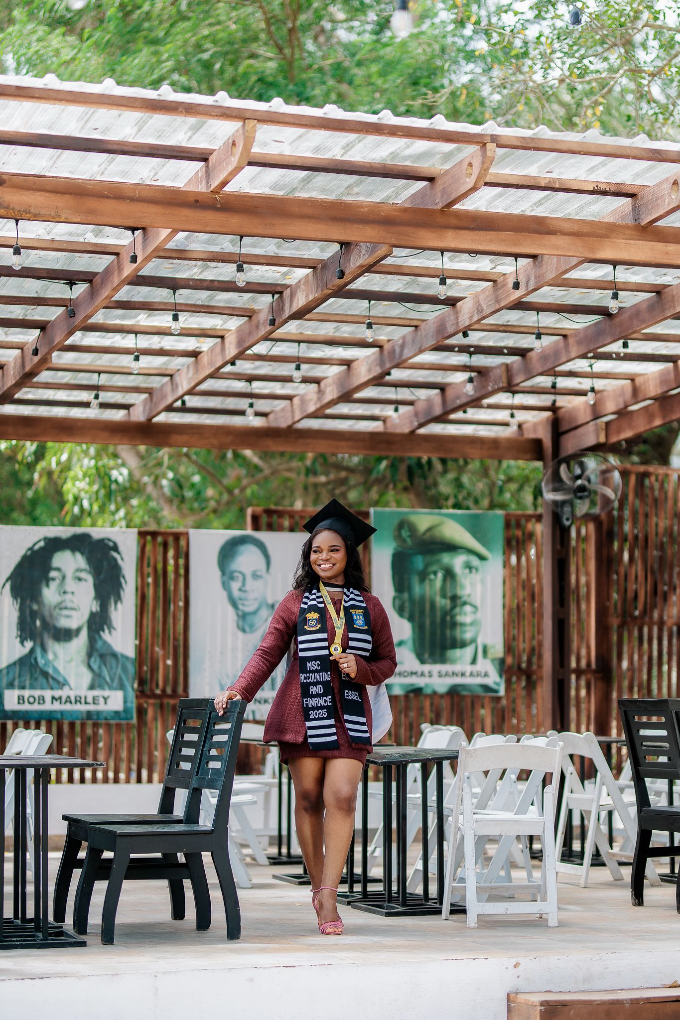 Smiling Black female graduate in cap and gown walking under wooden pergola near Bob Marley mural.