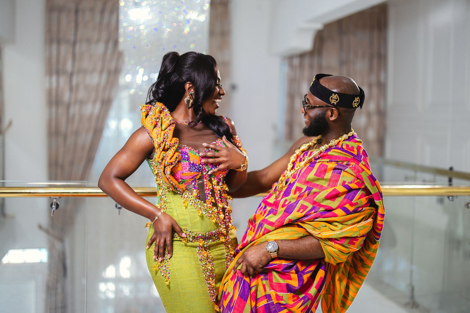 A couple poses in vibrant Kente cloth traditional Ghanaian wedding attire on a balcony.