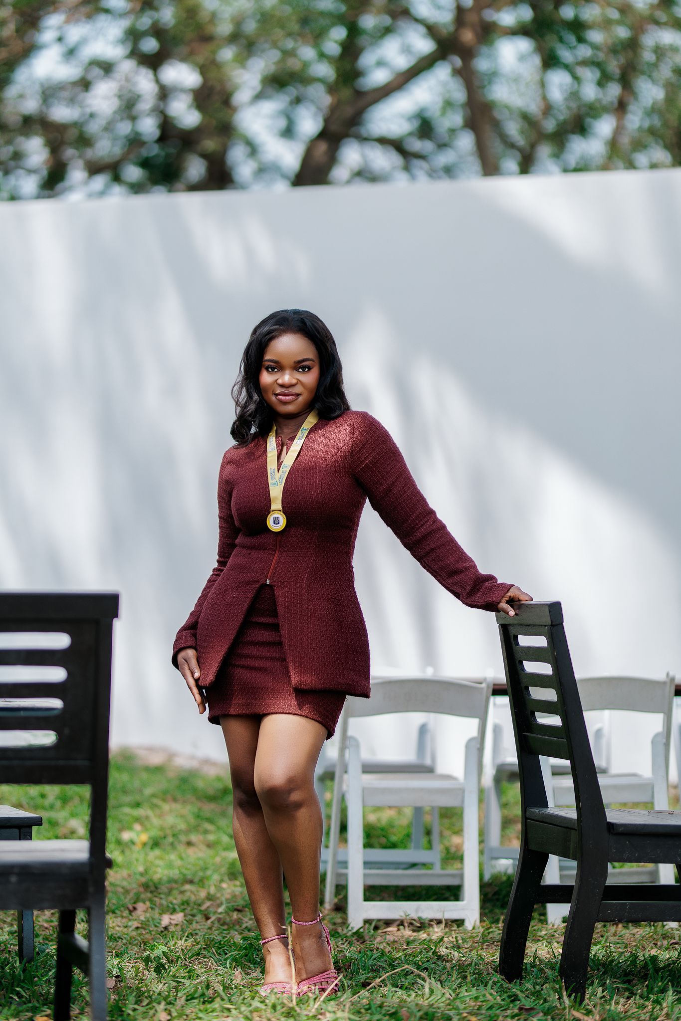 Young Black woman in burgundy mini dress with gold medal, posing outdoors between chairs.