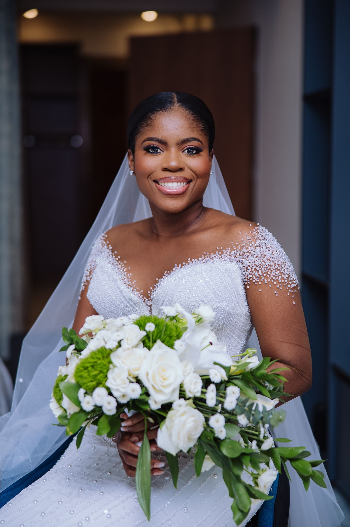 Radiant bride in beaded white wedding gown holds green and white floral bouquet with veil.