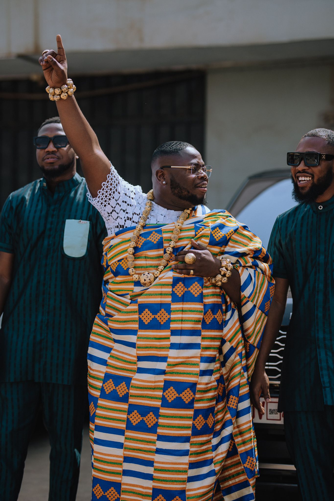 A groom in traditional Kente cloth raises his hand while groomsmen in teal outfits stand beside him.