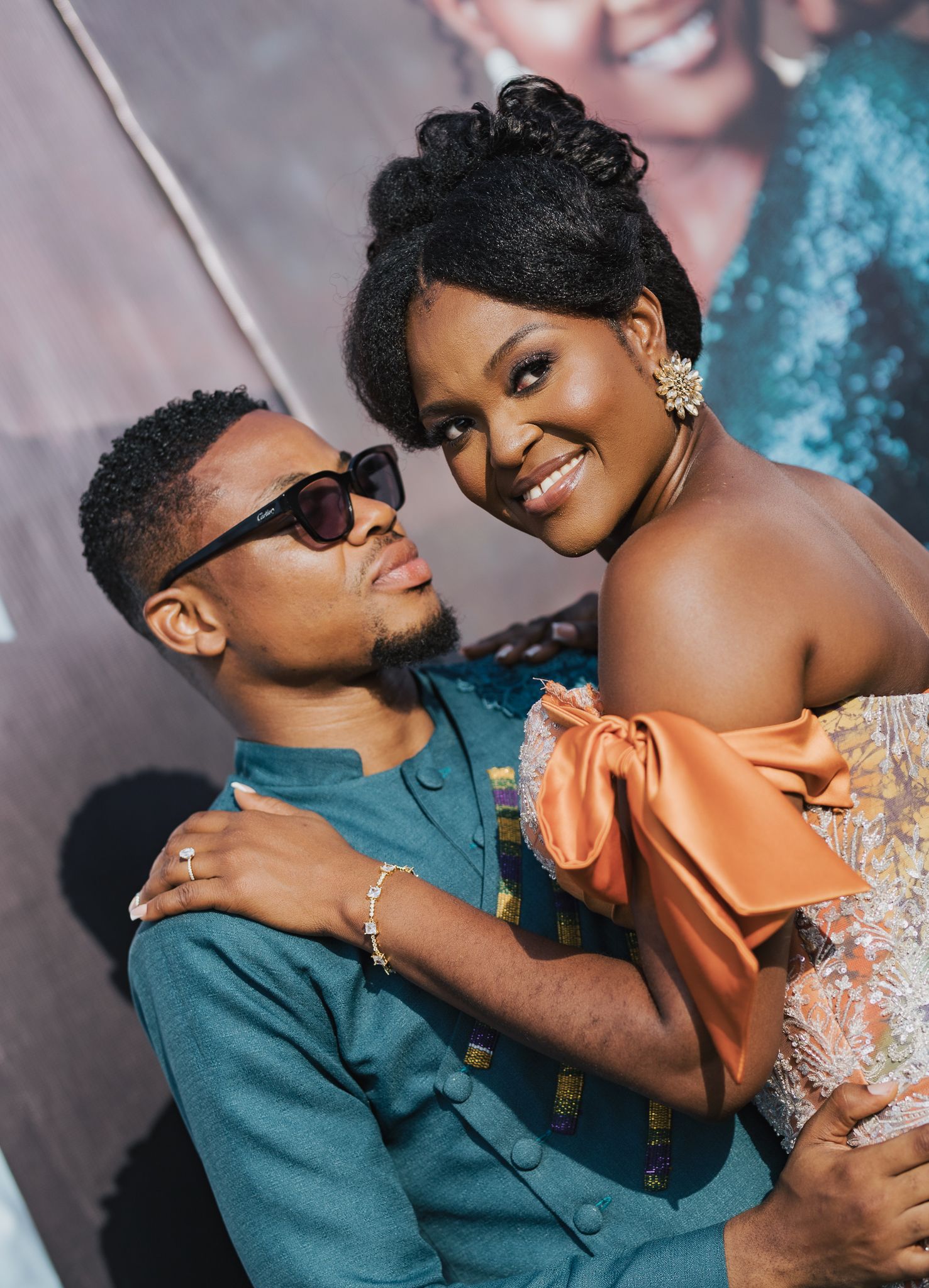 Couple in elegant African attire embrace warmly at a formal event, woman in orange bow dress smiles joyfully.