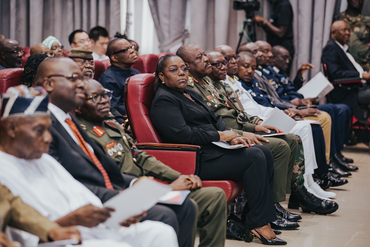 Diverse audience of military and civilian officials seated in red chairs at a formal conference event.