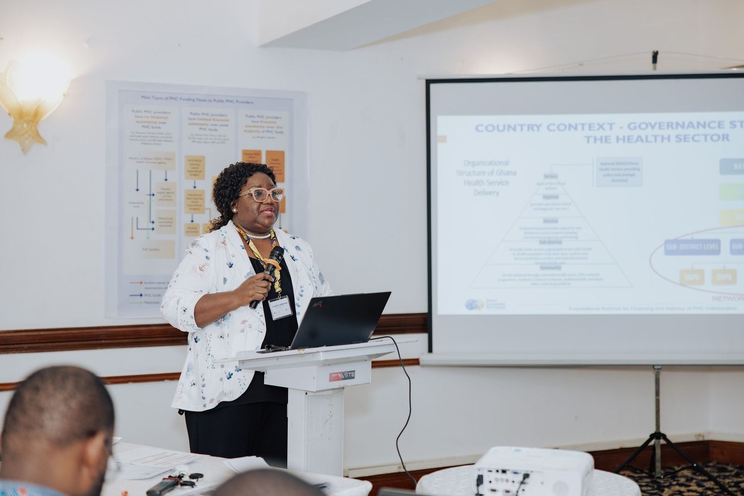 Woman presenting at a conference, standing at a podium with a laptop, gesturing toward a projected slide on governance.