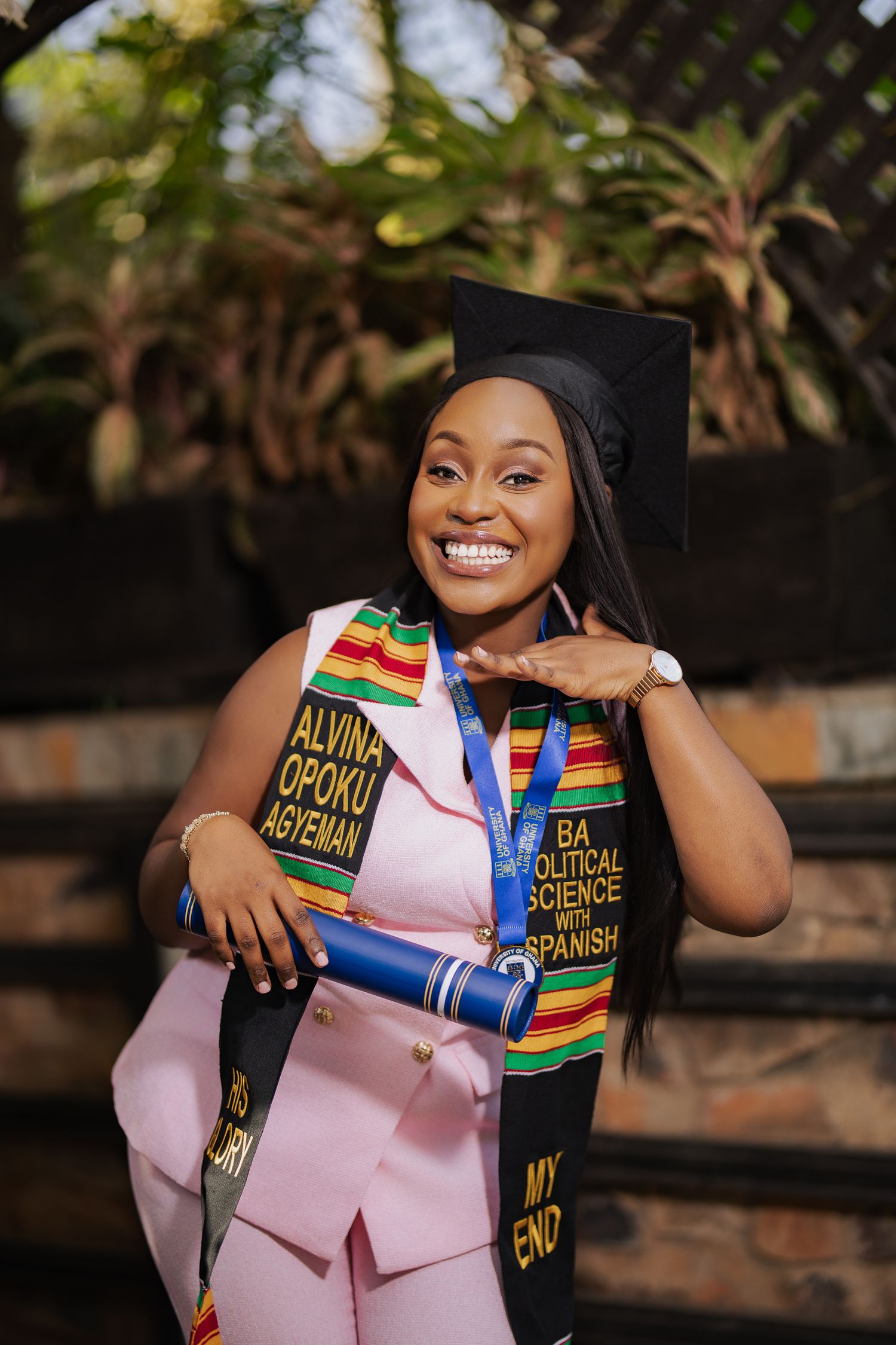 Smiling graduate Alvina Opoku Agyeman in pink suit with kente stole and Political Science Spanish degree.
