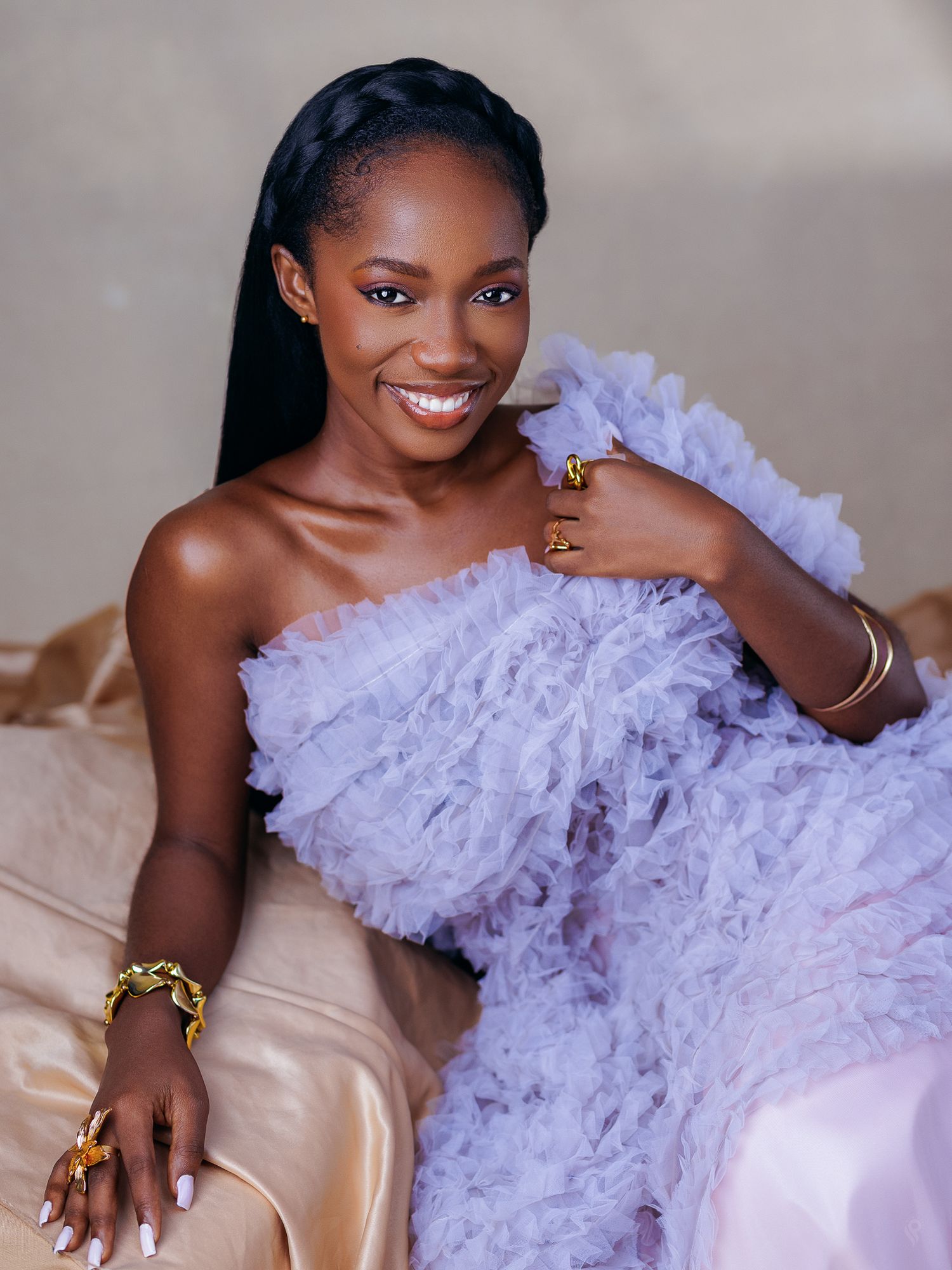 Smiling woman in lavender ruffled off-shoulder gown poses with gold jewelry accessories.