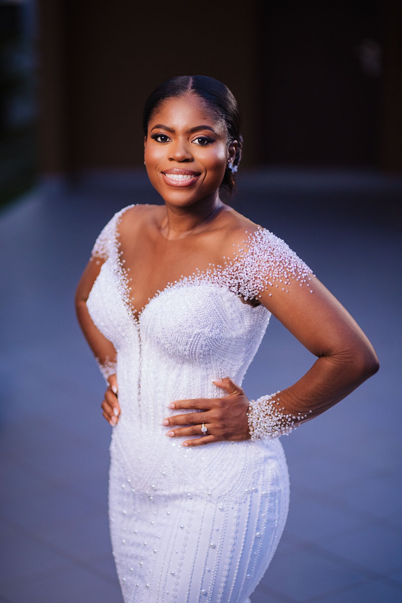 Smiling bride in elegant white beaded wedding gown posing outdoors at dusk.
