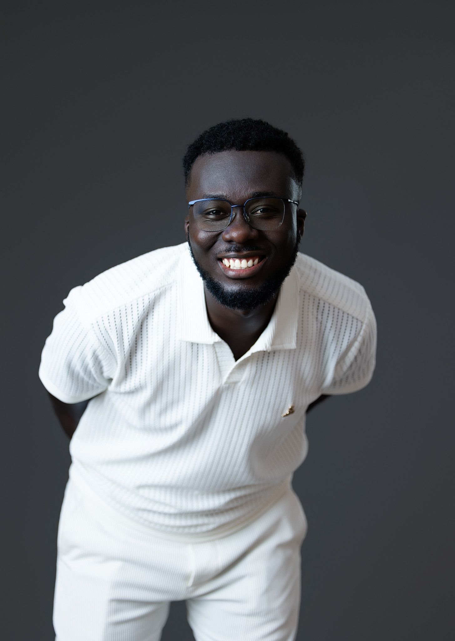 Joyful Black man in white polo shirt and glasses leaning forward smiling against gray background.