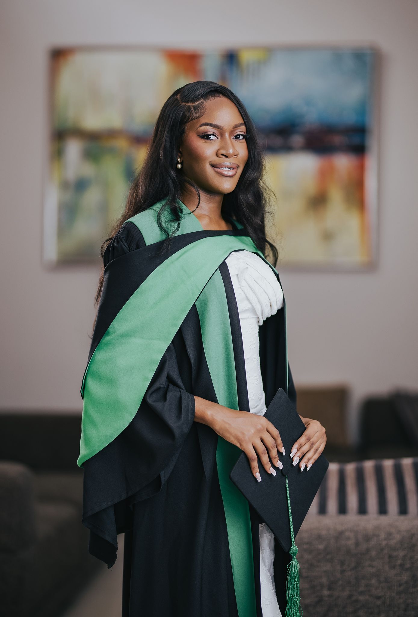 Beautiful graduate in black and green academic regalia holding cap while smiling in elegant indoor setting.