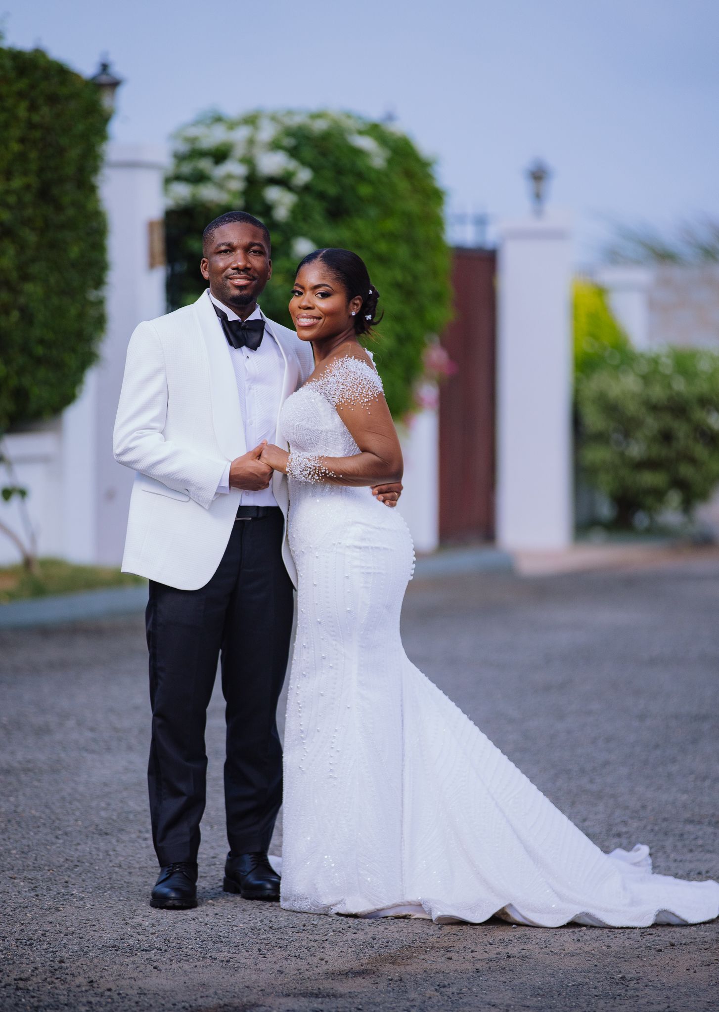 Elegant couple posing in wedding attire, groom in white tuxedo and bride in beaded mermaid gown outdoors.