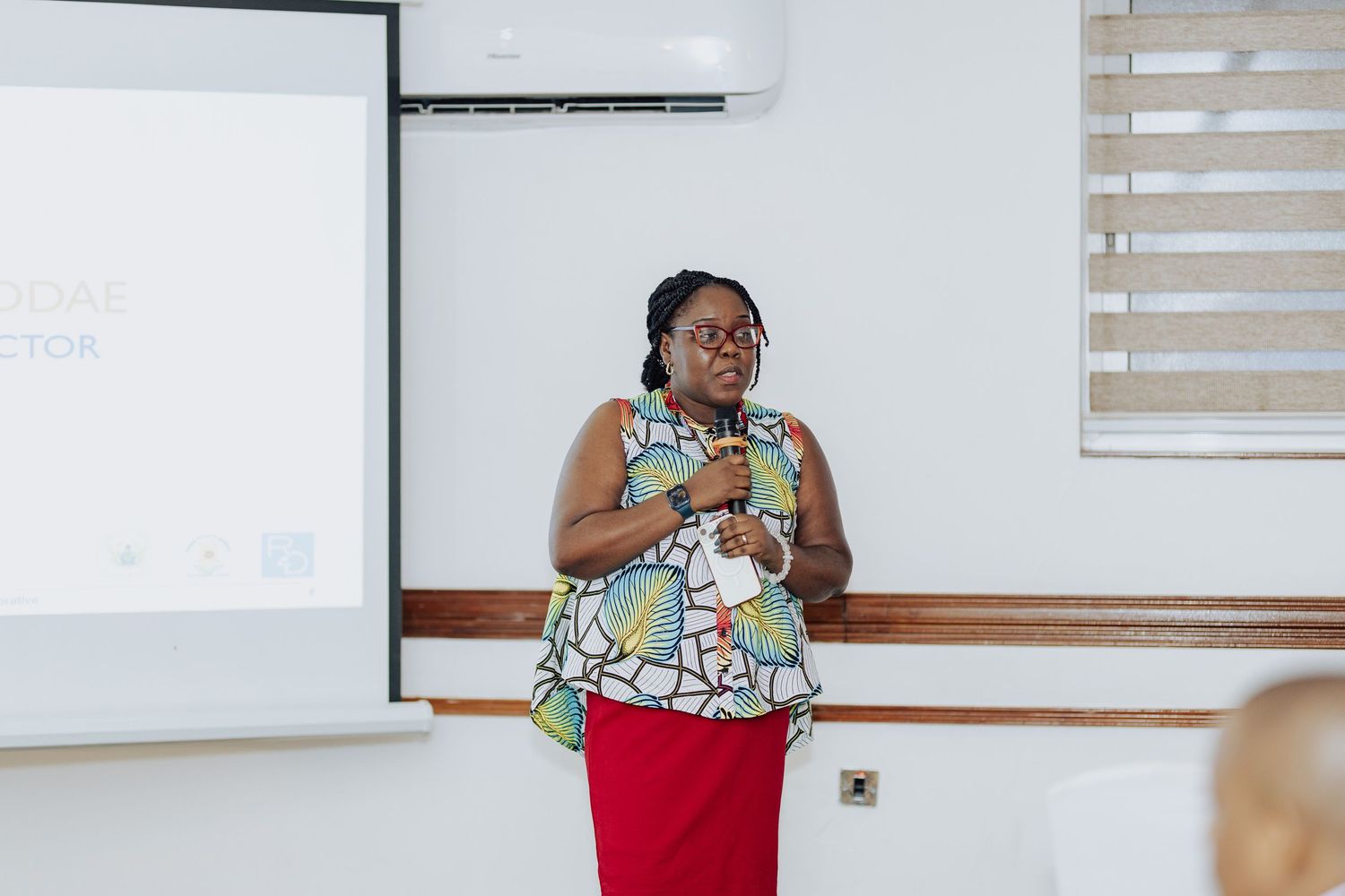 Woman in floral top and red skirt presenting at a whiteboard during a professional seminar or conference.