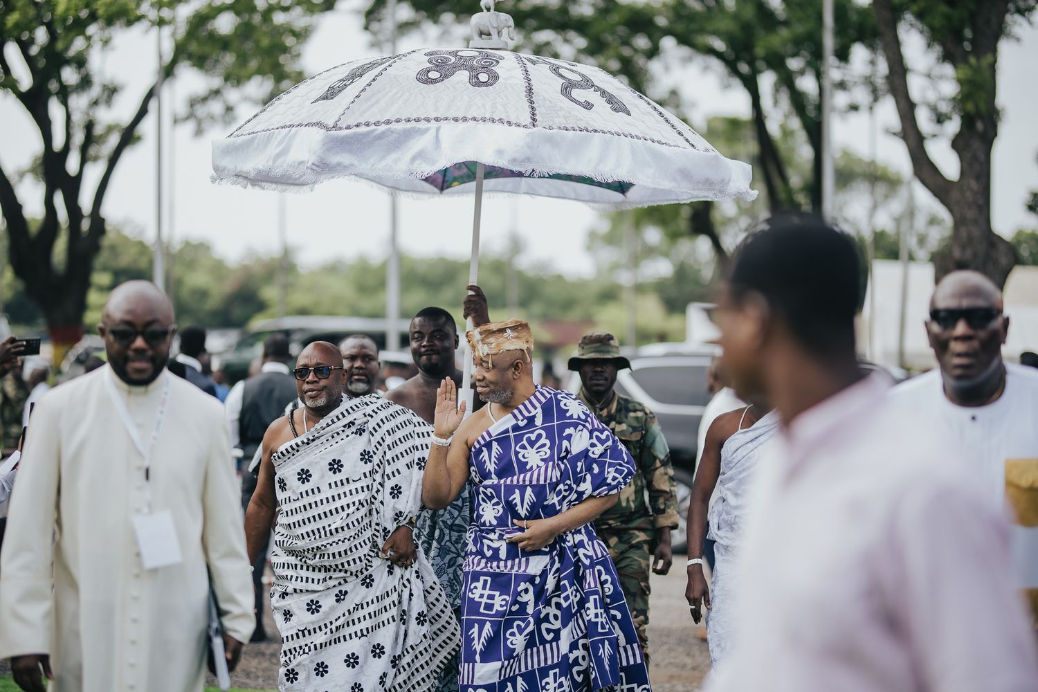 Ghanaian chief in traditional kente cloth walking under ceremonial umbrella outdoors.
