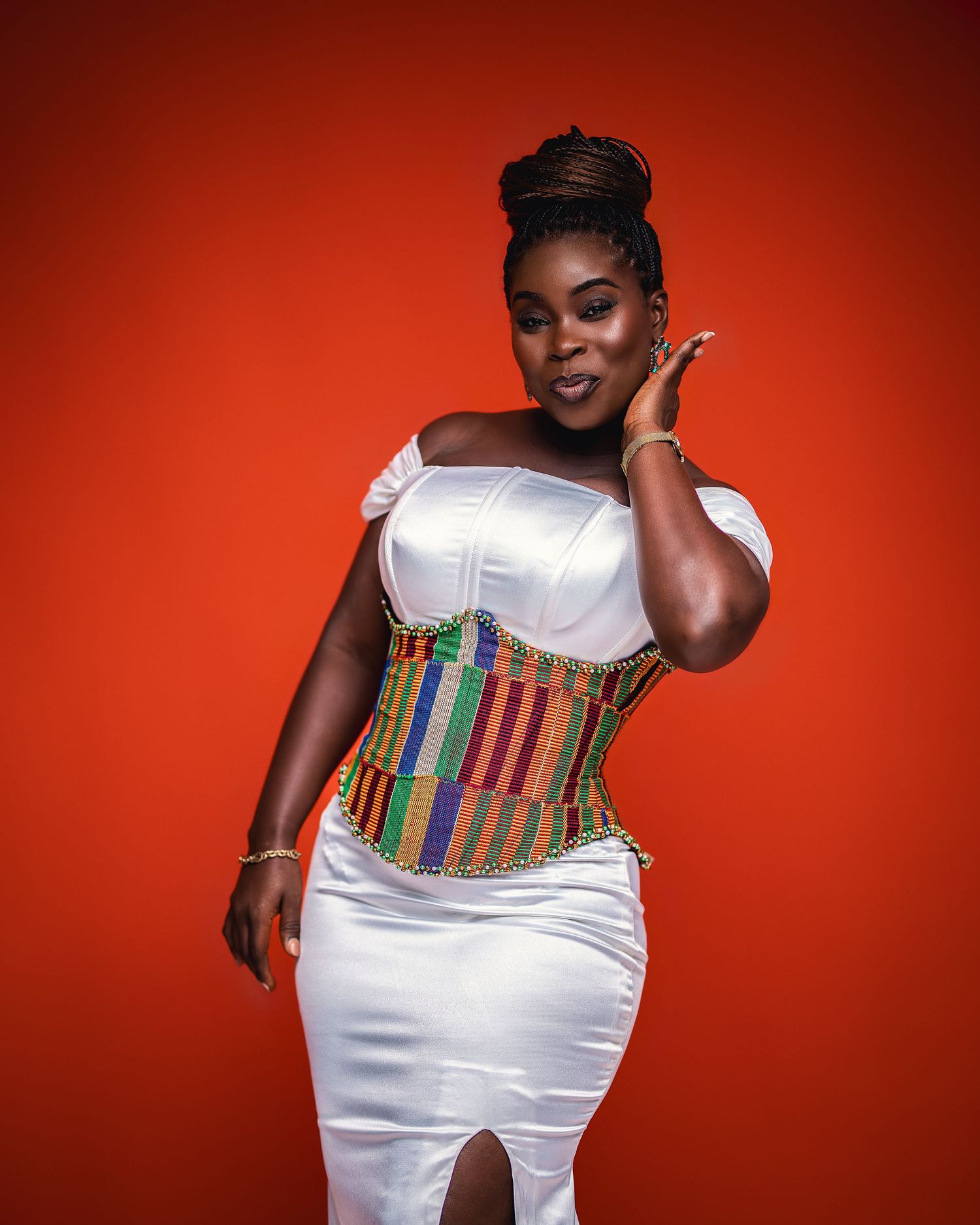 Woman in white dress with colorful kente corset posing against vibrant red background.