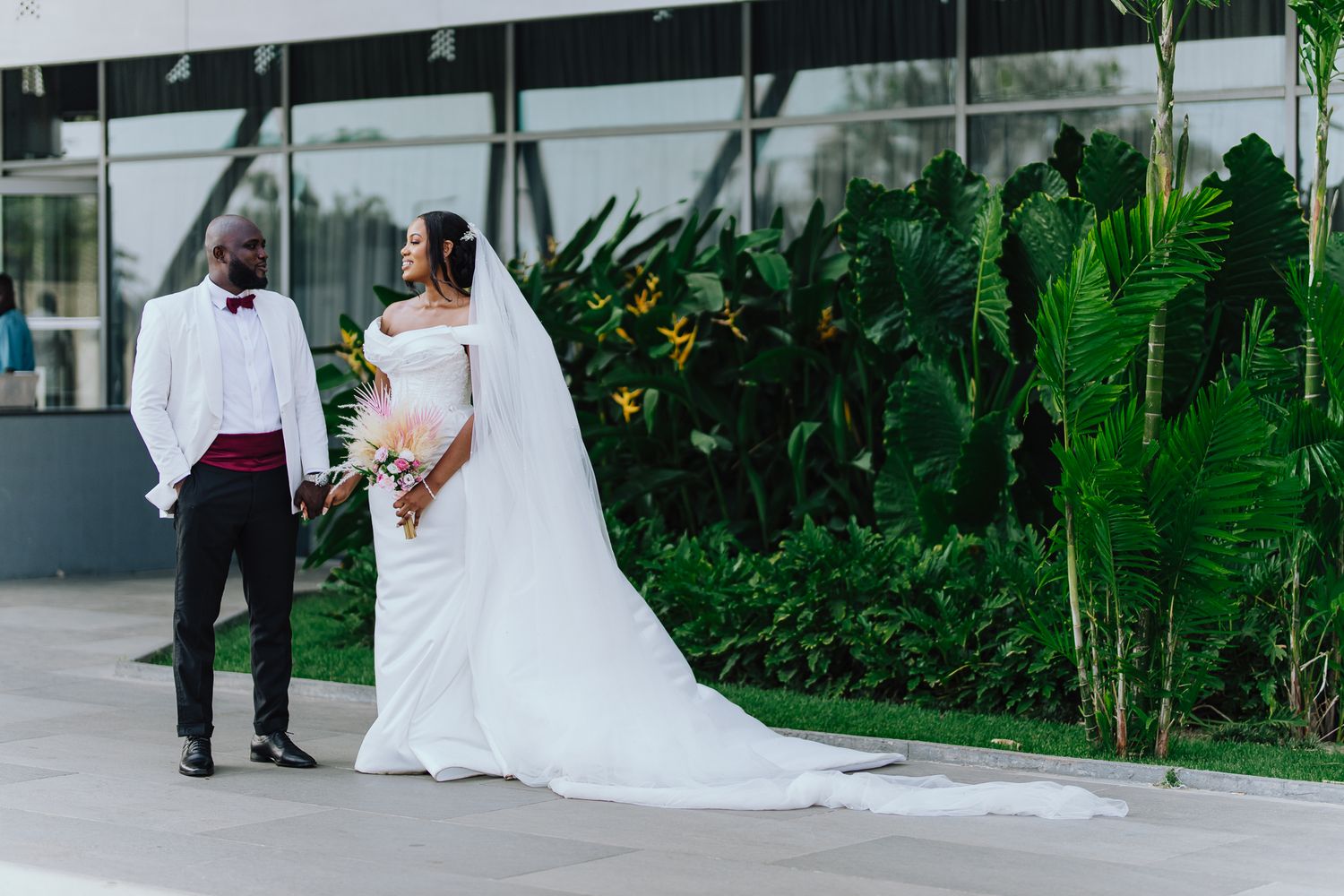 Bride and groom in white wedding attire posing before lush tropical greenery outdoors.