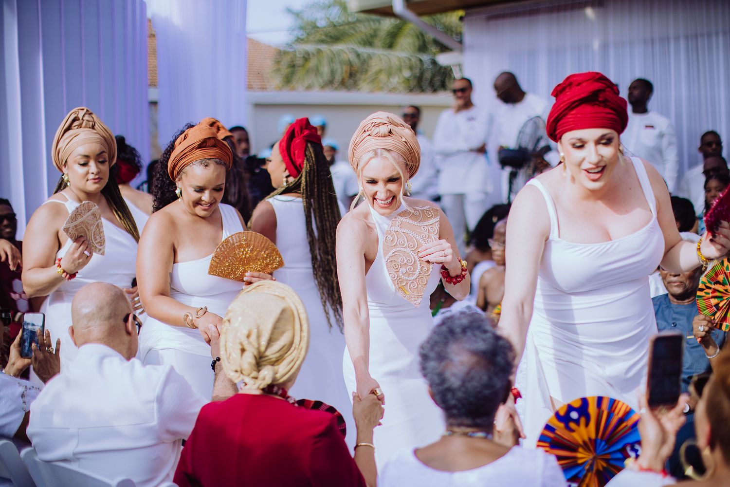Bridesmaids in white dresses and colorful head wraps holding fans during traditional African wedding ceremony.