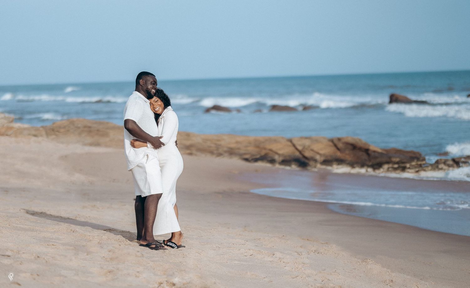 Couple in matching white outfits embracing romantically on a sandy beach by the ocean.