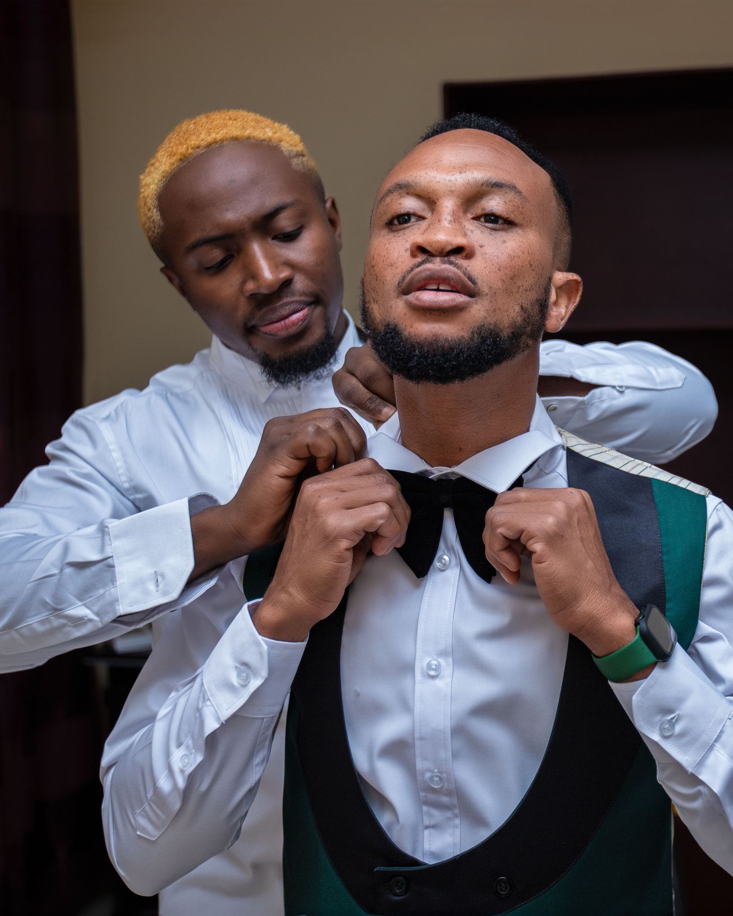 A man helps a groom in a vest adjust his bow tie during wedding preparations.