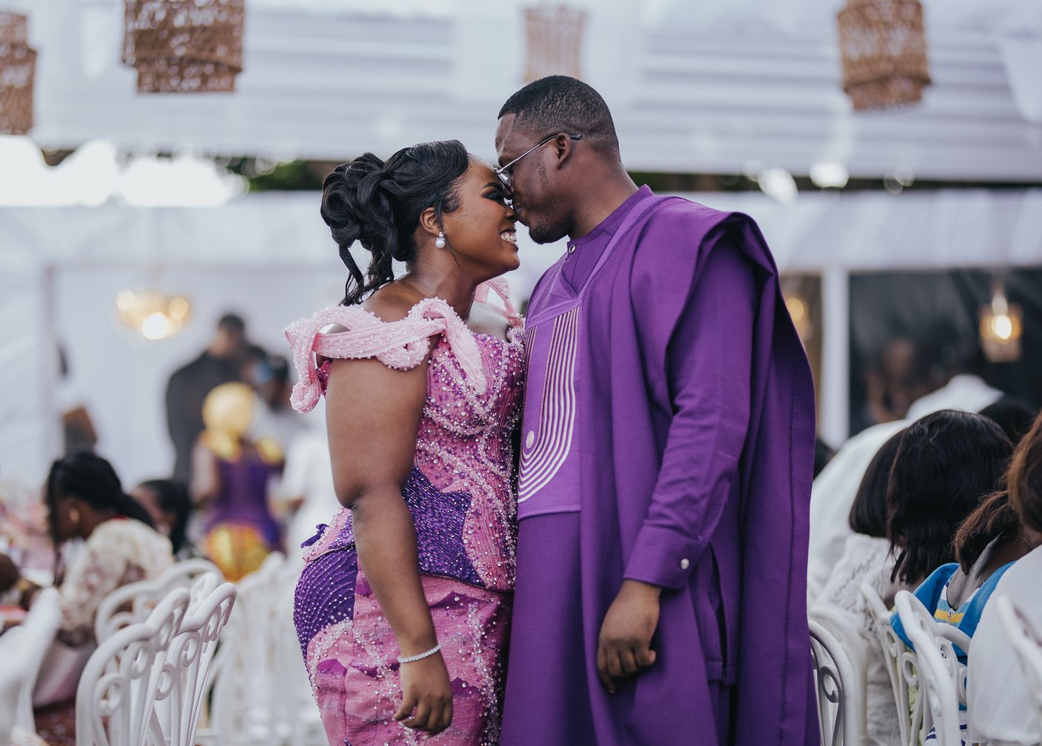 Romantic couple in purple and pink African attire share an intimate moment at a wedding reception.