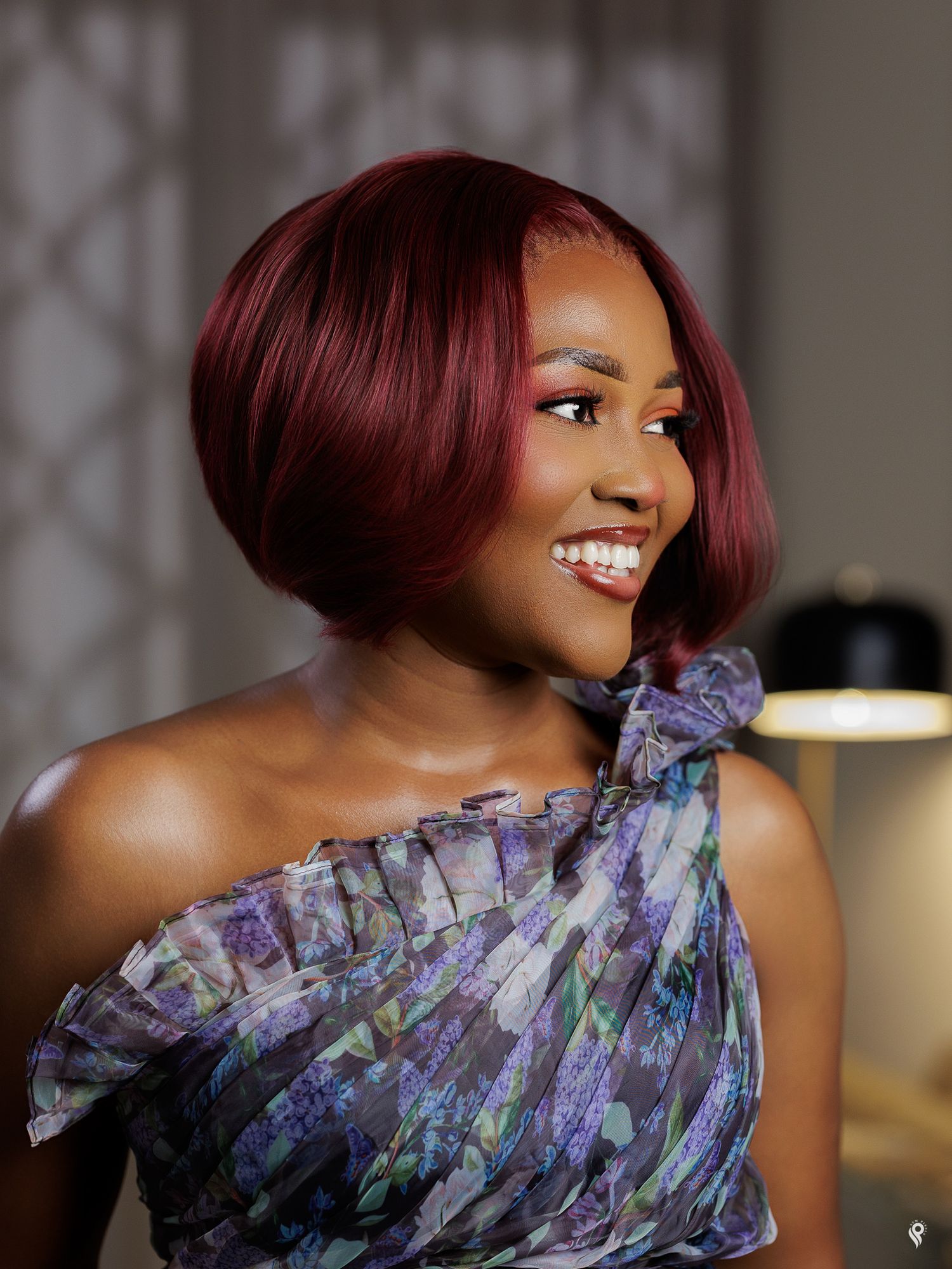 Glamorous Black woman with burgundy bob hairstyle in floral one-shoulder dress, smiling in soft light.