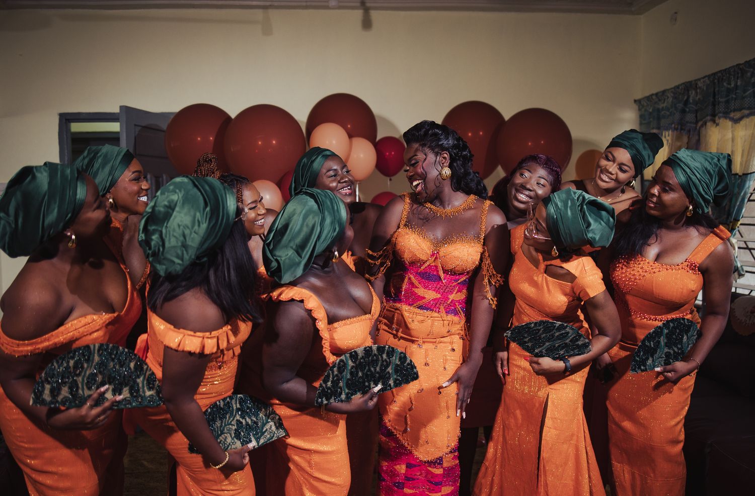 Bride with bridesmaids in orange dresses and green headwraps celebrating at traditional Ghanaian wedding.