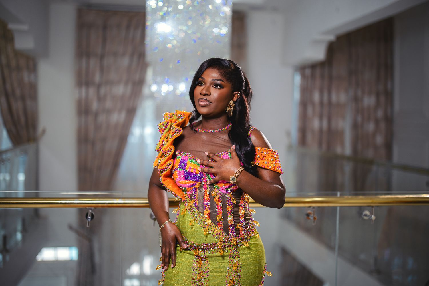 A woman in a colorful orange and purple traditional African outfit poses on a balcony.