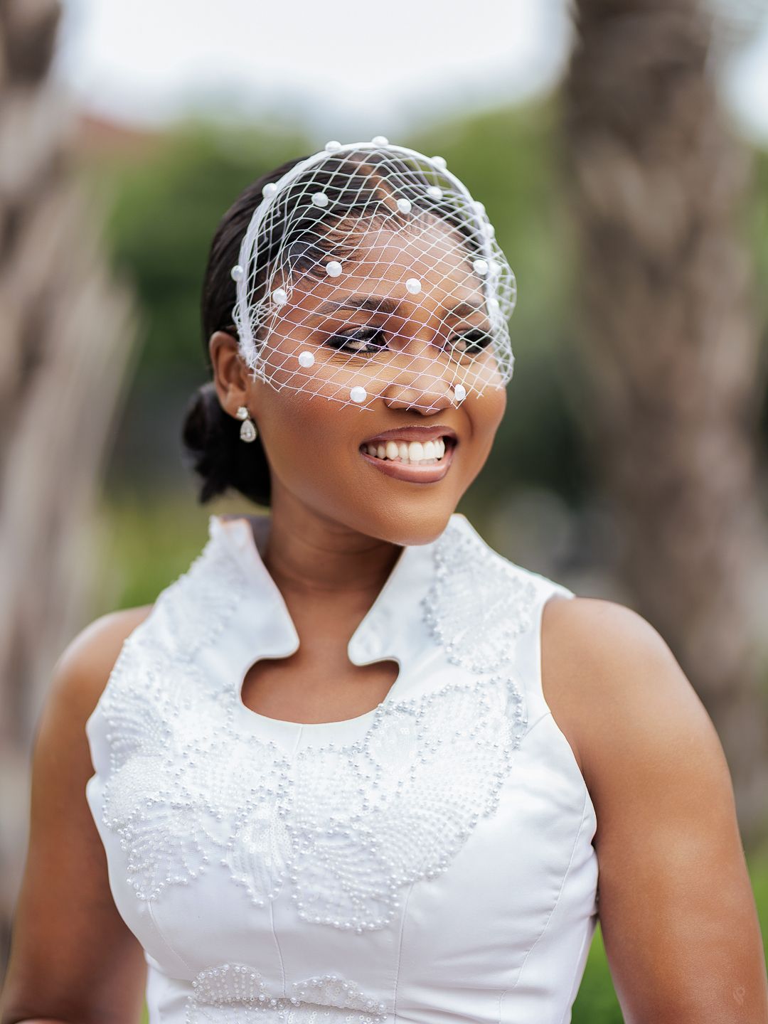 A smiling bride wearing a delicate pearl-adorned birdcage veil and white lace wedding dress outdoors.