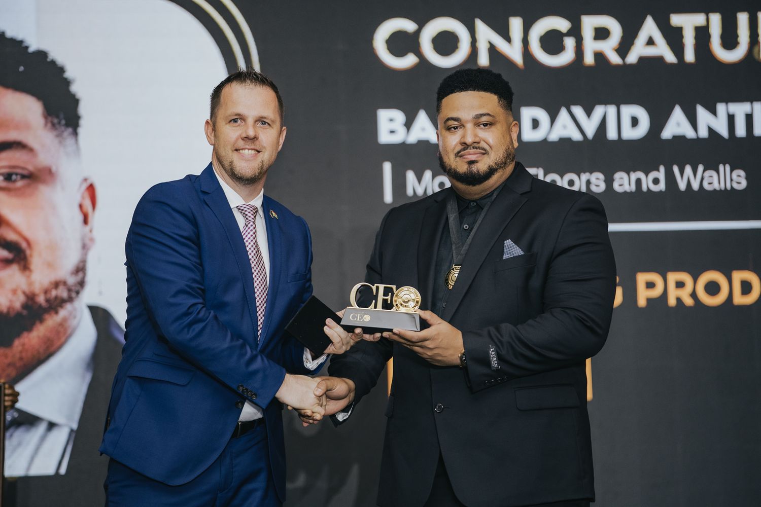 Two men in formal attire shake hands while presenting a congratulatory award at ceremony.