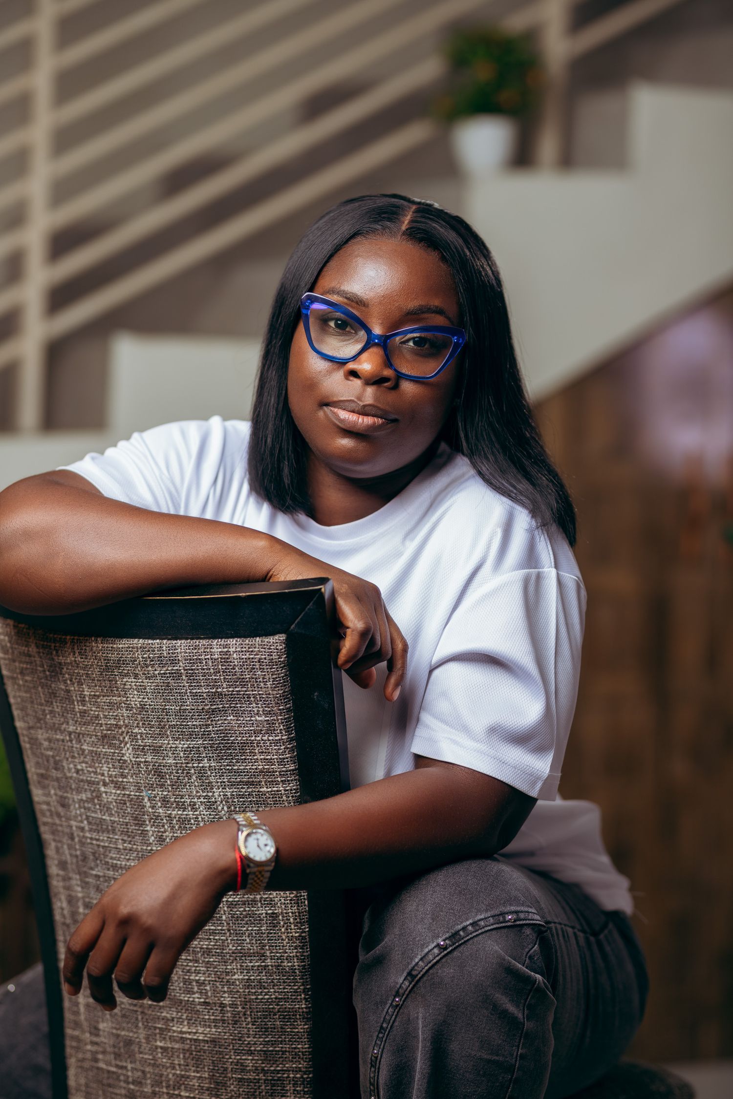 A young Black woman in a white t-shirt and blue cat-eye glasses sits relaxed in a chair, wearing a watch.
